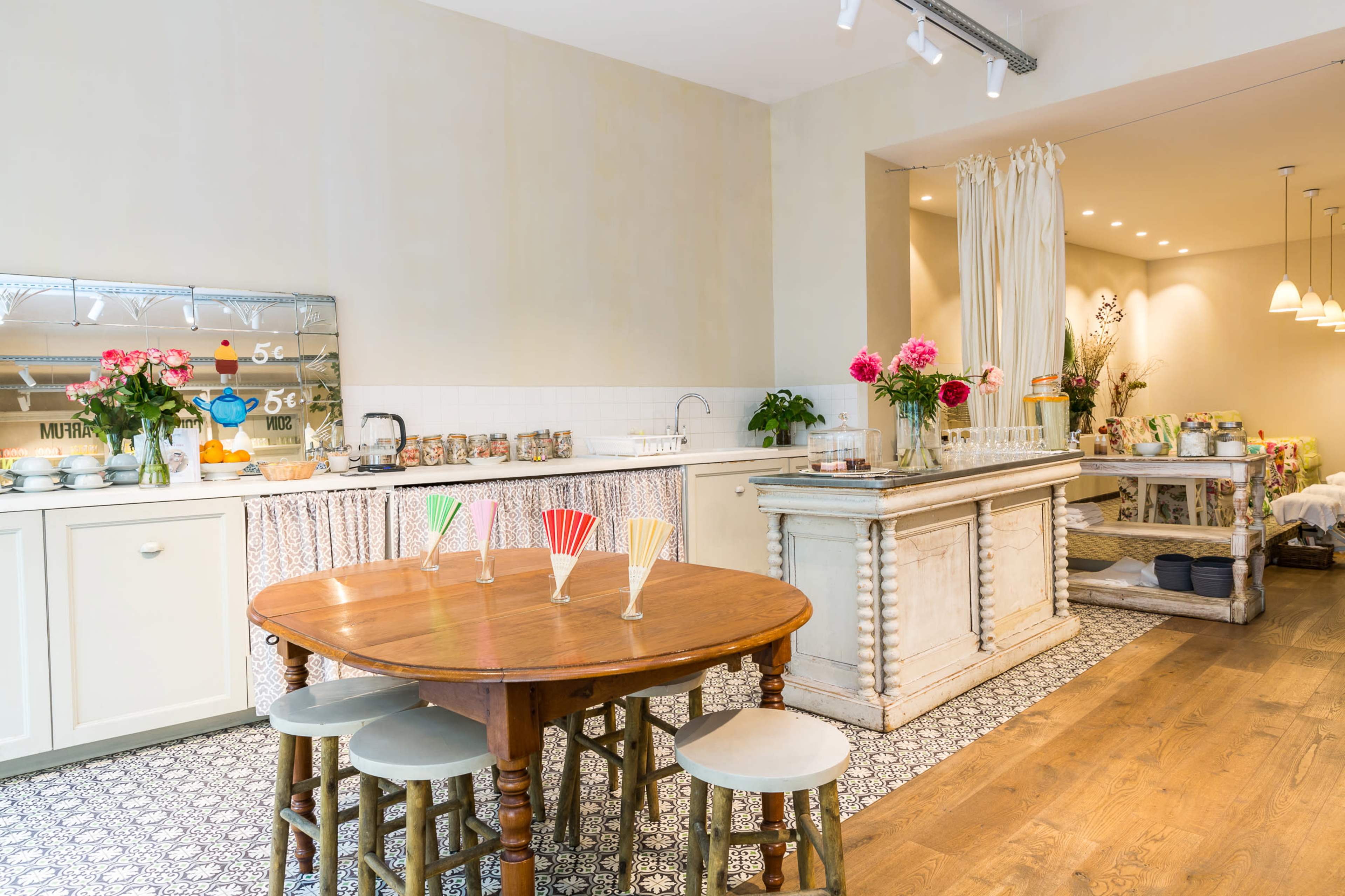 A bright, airy kitchen and dining area with a wooden table, pastel-colored chairs, and a counter lined with jars and flowers.