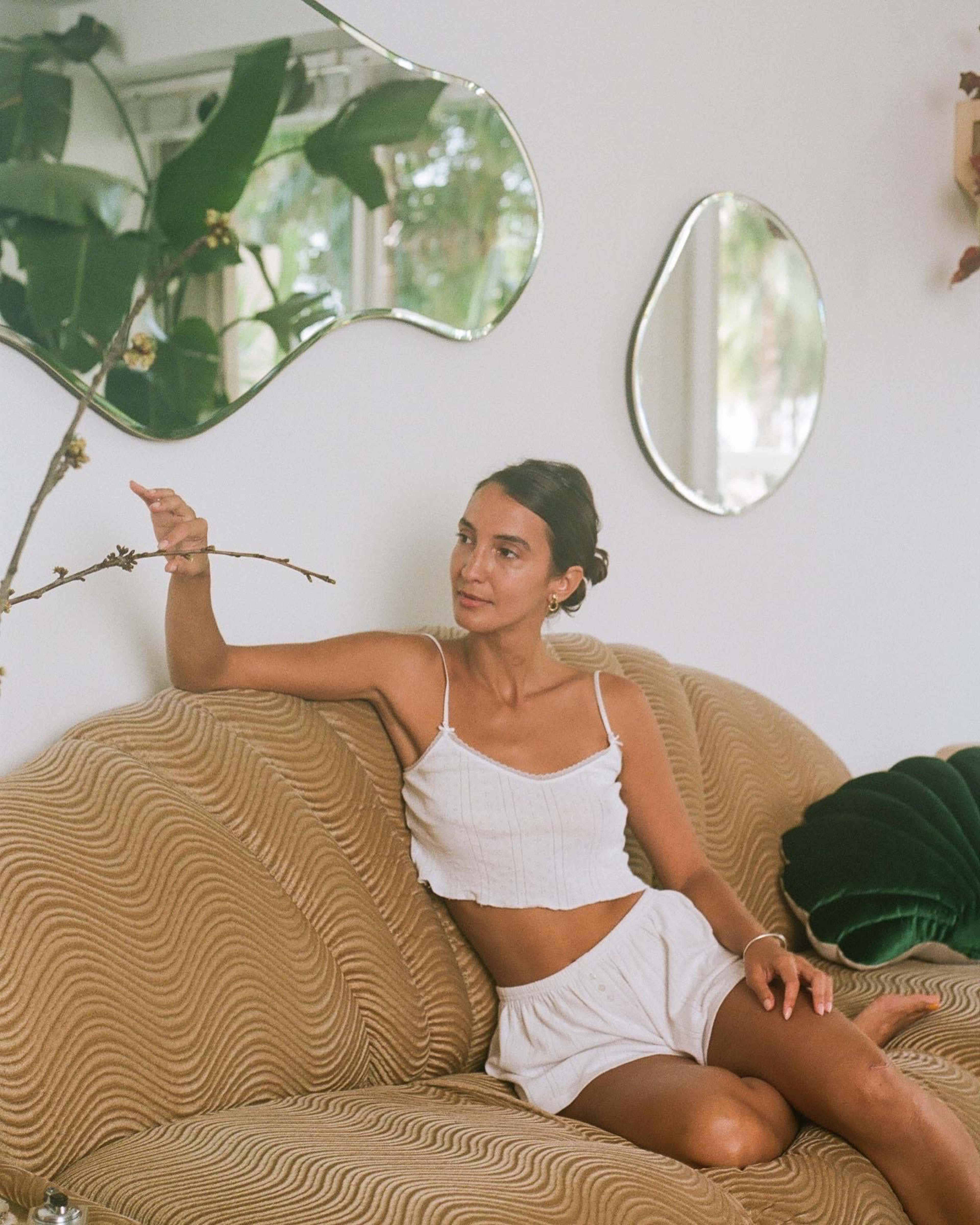 A woman sitting on a wavy-patterned couch points towards a branch while looking at her reflection in a nearby mirror.