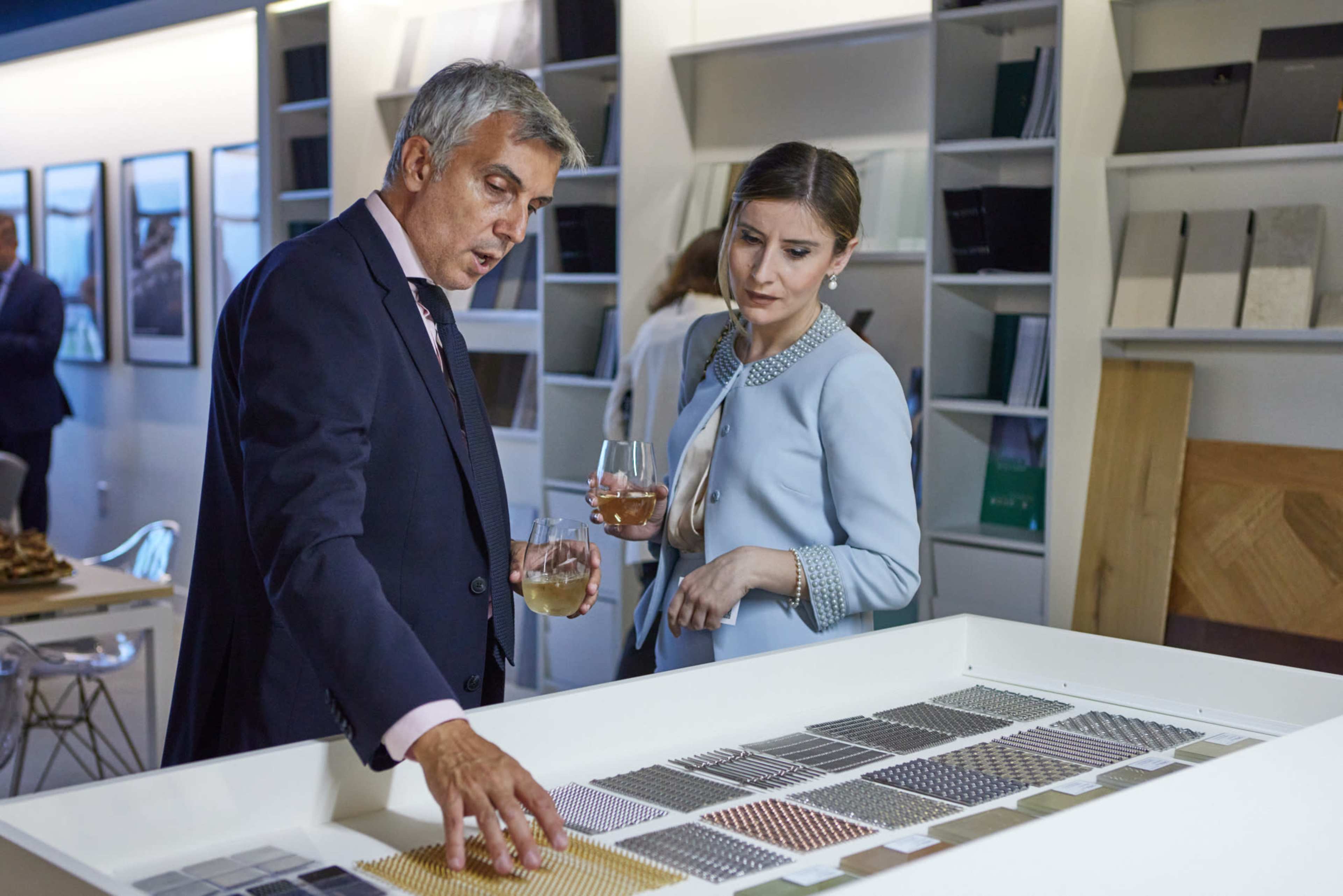 A man and a woman stand by a display of material samples, discussing options while holding glasses of drinks.