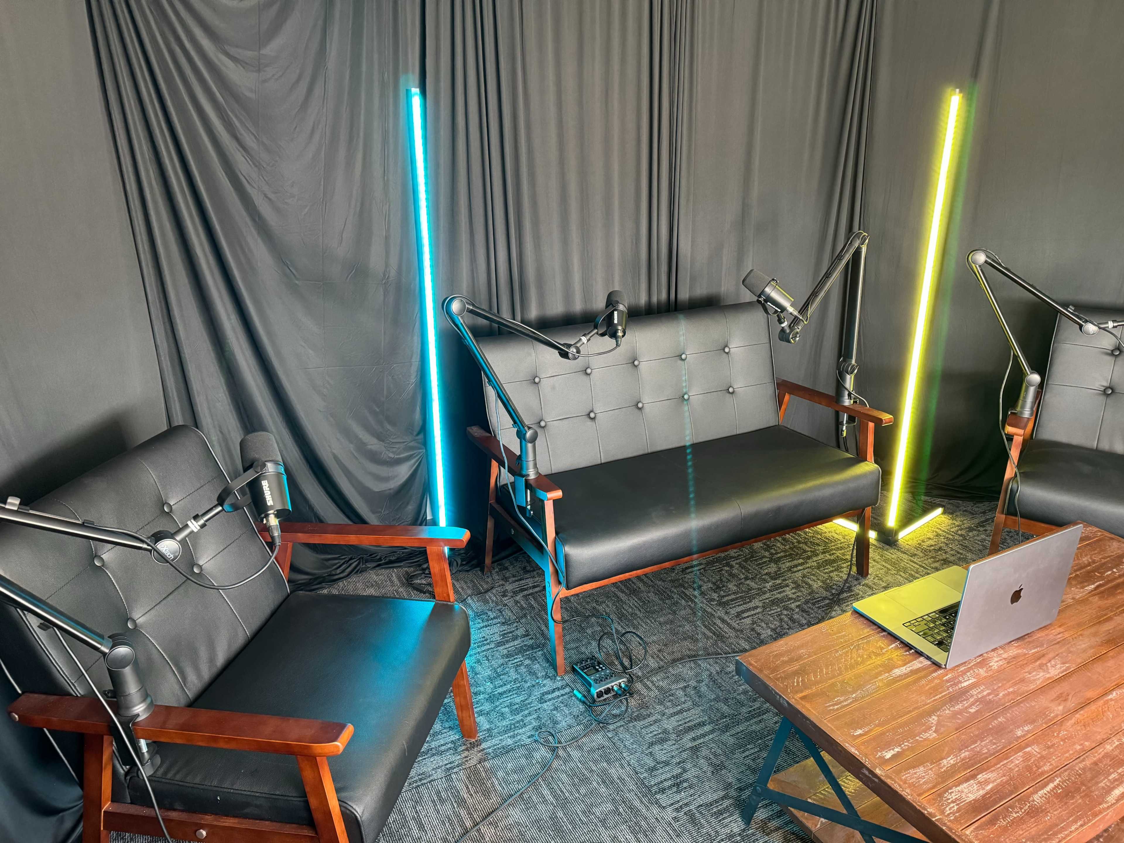 A small studio with two black couches, each equipped with a microphone on an arm, and illuminated by vertical neon light poles on either side, along with a laptop on a wooden table in the center.