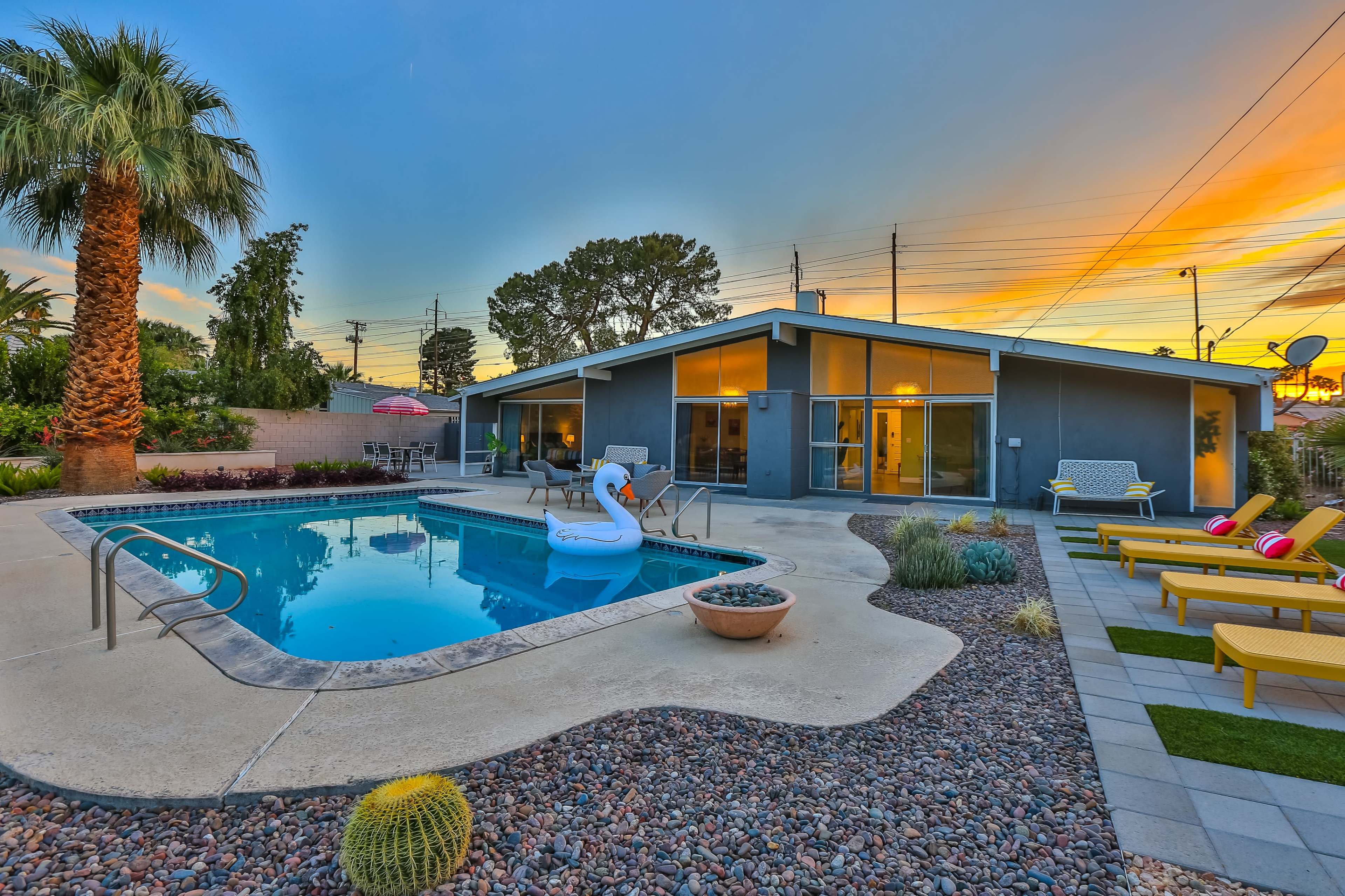 A modern backyard features a swimming pool with a swan float, surrounded by lounge chairs and palm trees, set against a vibrant sunset.