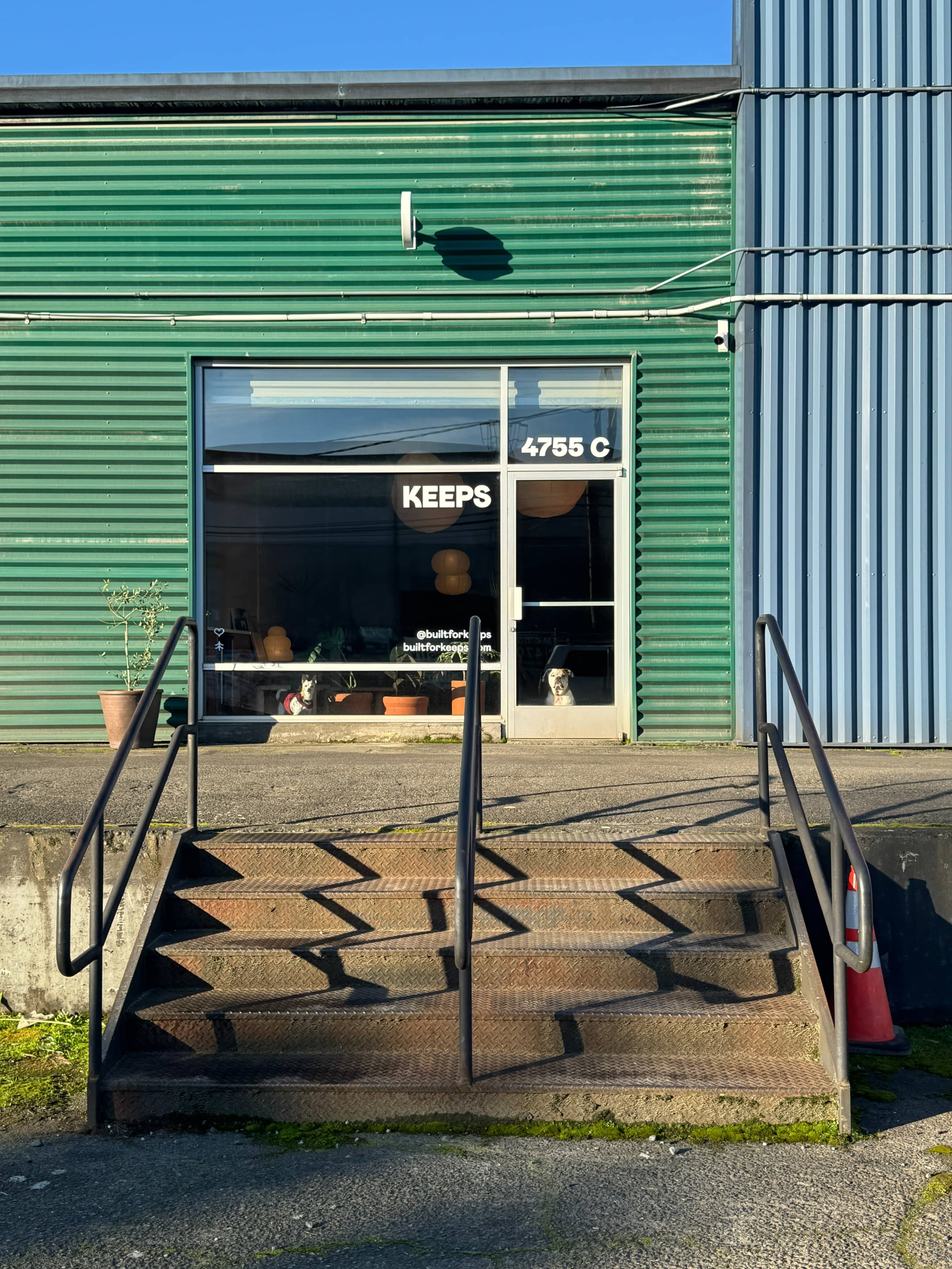 A set of metal stairs leads up to a storefront with a large window displaying the word "KEEPS" and a glimpse of interior elements.