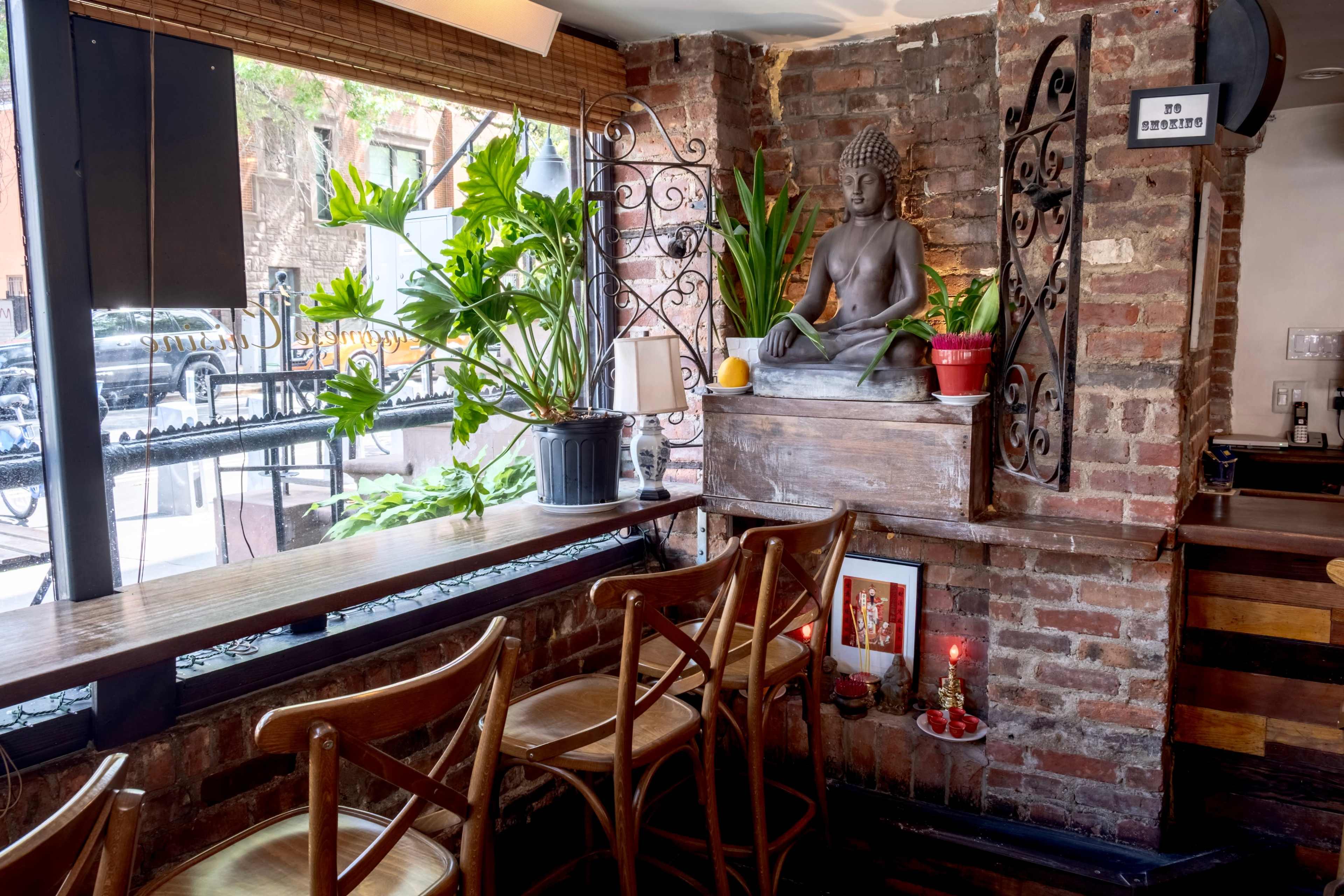 The image shows a cozy café interior featuring wooden stools, a brick wall with a Buddha statue, and various indoor plants.
