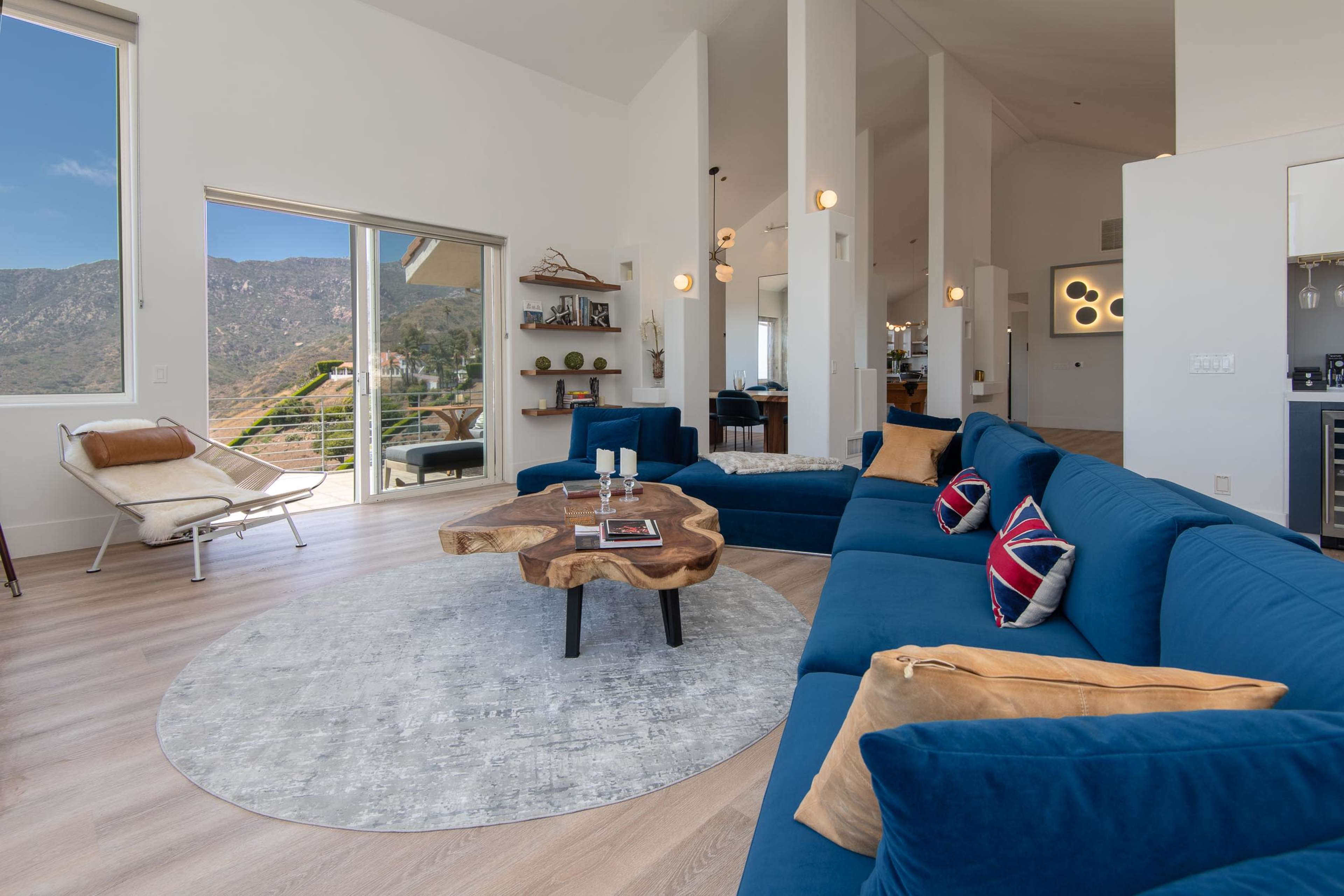 A spacious living room featuring a large blue sectional sofa, a wooden coffee table, and expansive windows overlooking a mountainous landscape.