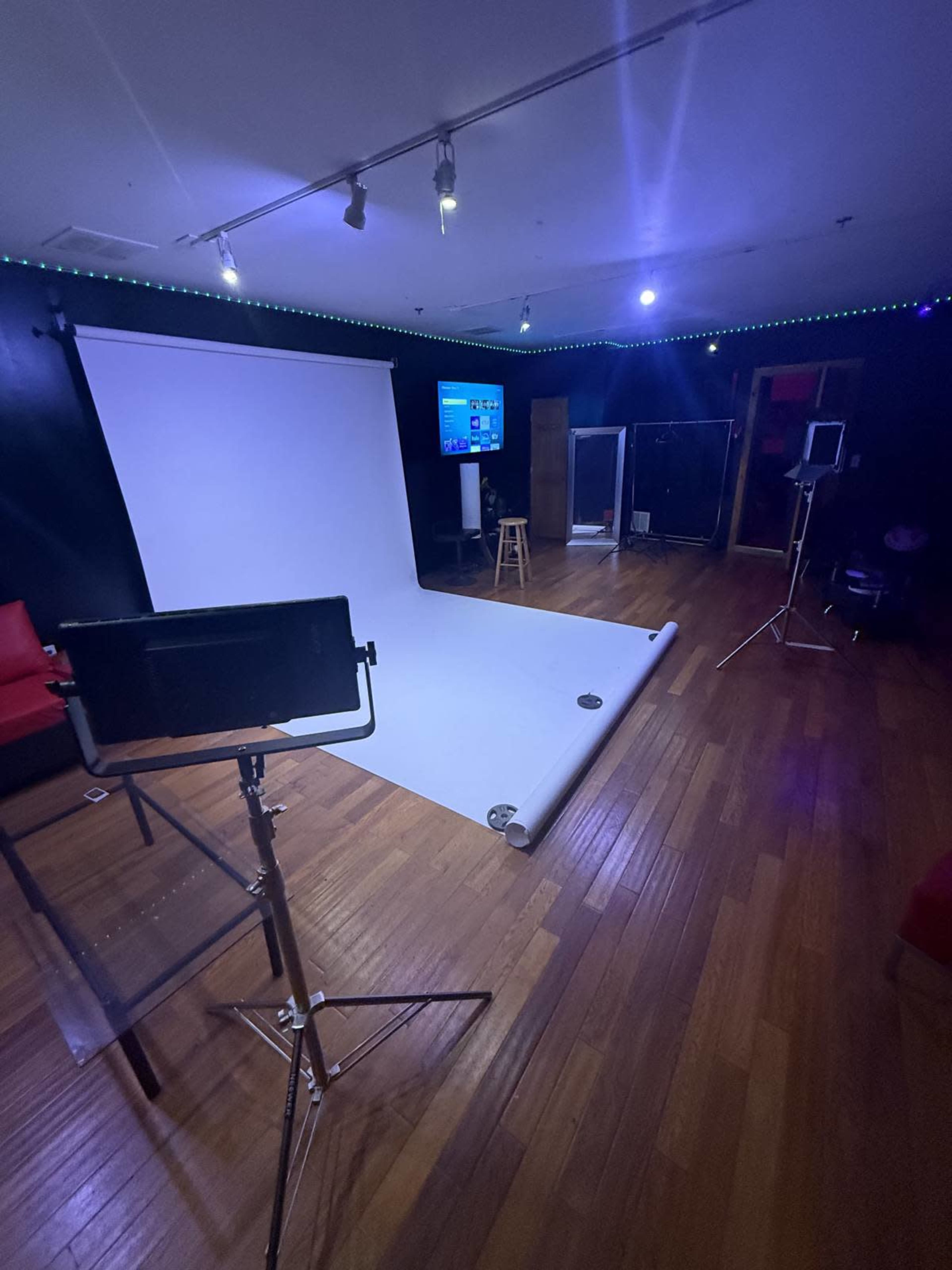 The image shows a photography studio with a white backdrop, a wooden floor, various lighting equipment, and a television on a wall.