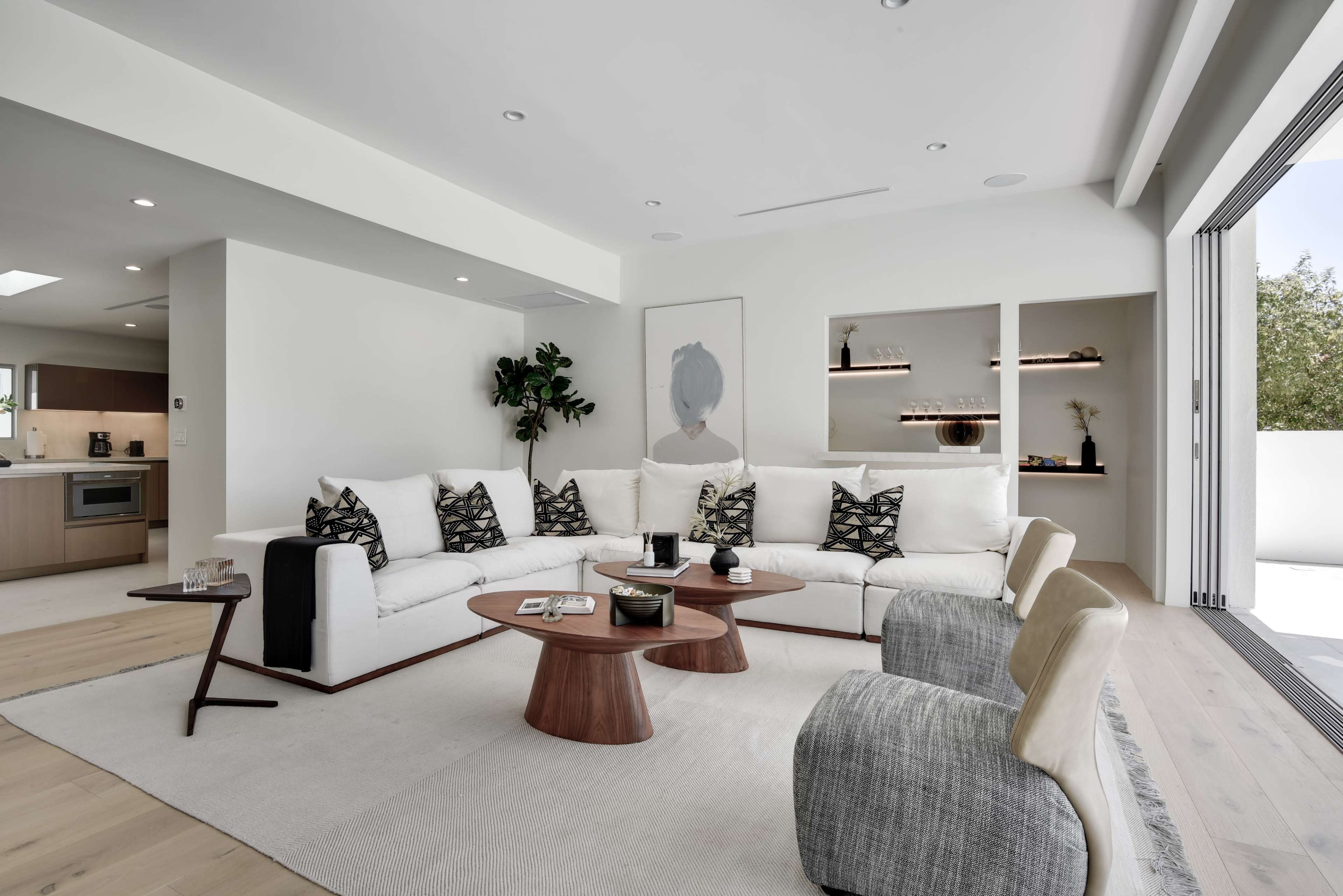 A modern living room features a large sectional sofa, a round wooden coffee table, and a light-colored area rug, with large windows offering natural light.