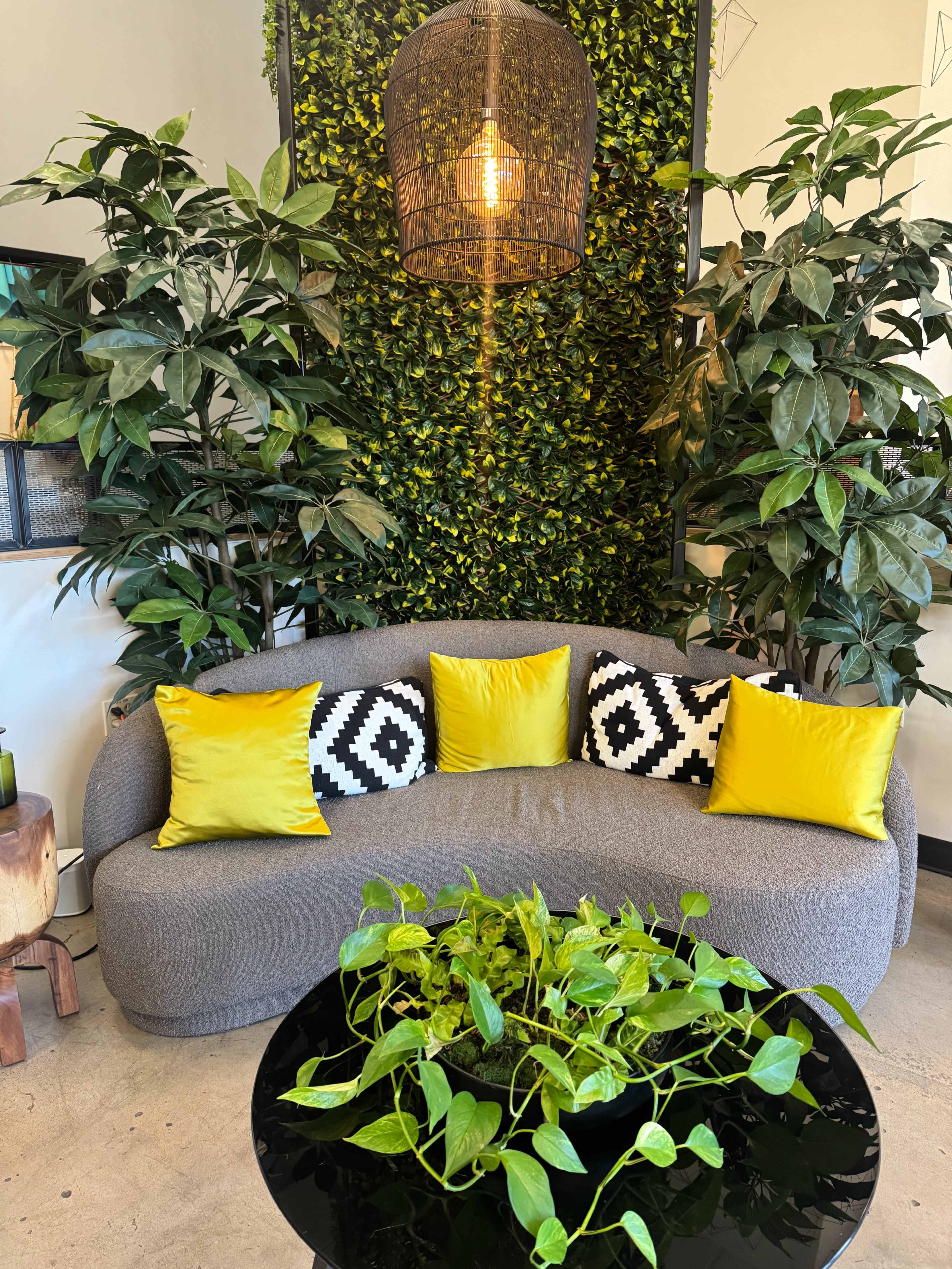 A curved gray sofa with yellow and patterned pillows is positioned in front of a green wall adorned with plants, accompanied by a hanging light fixture and a black table with a potted plant.