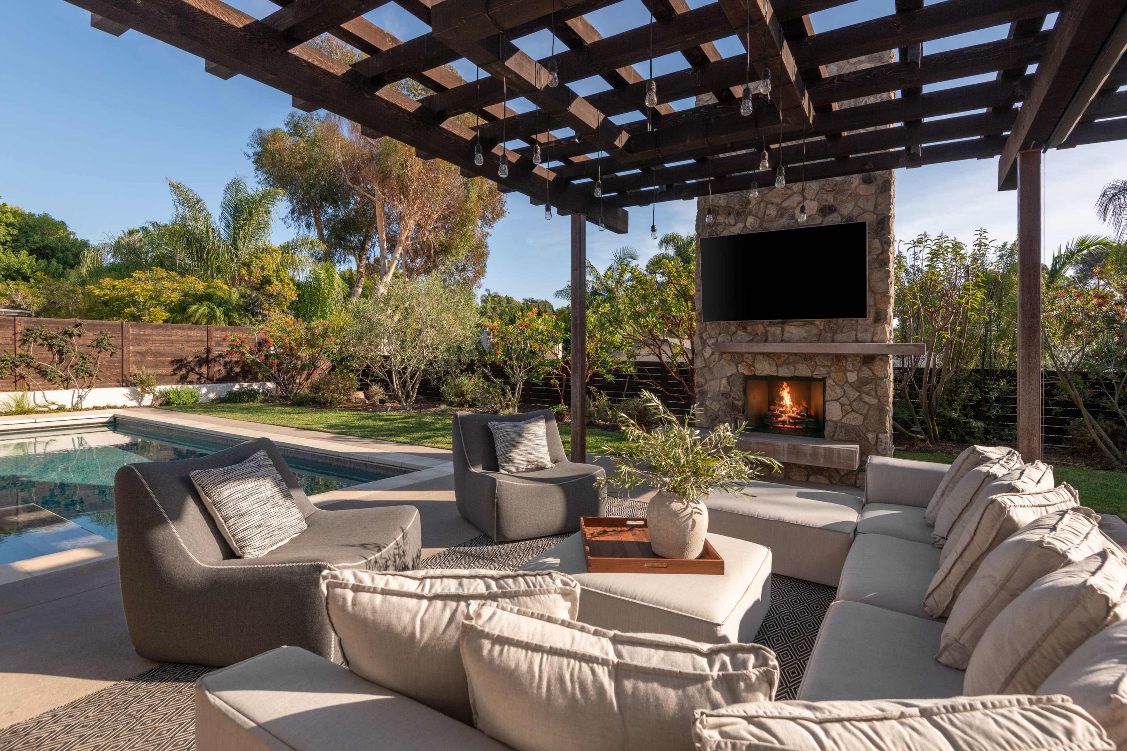 A modern outdoor living space features a stone fireplace, comfortable seating arranged in a circular layout, and a pool surrounded by lush greenery.