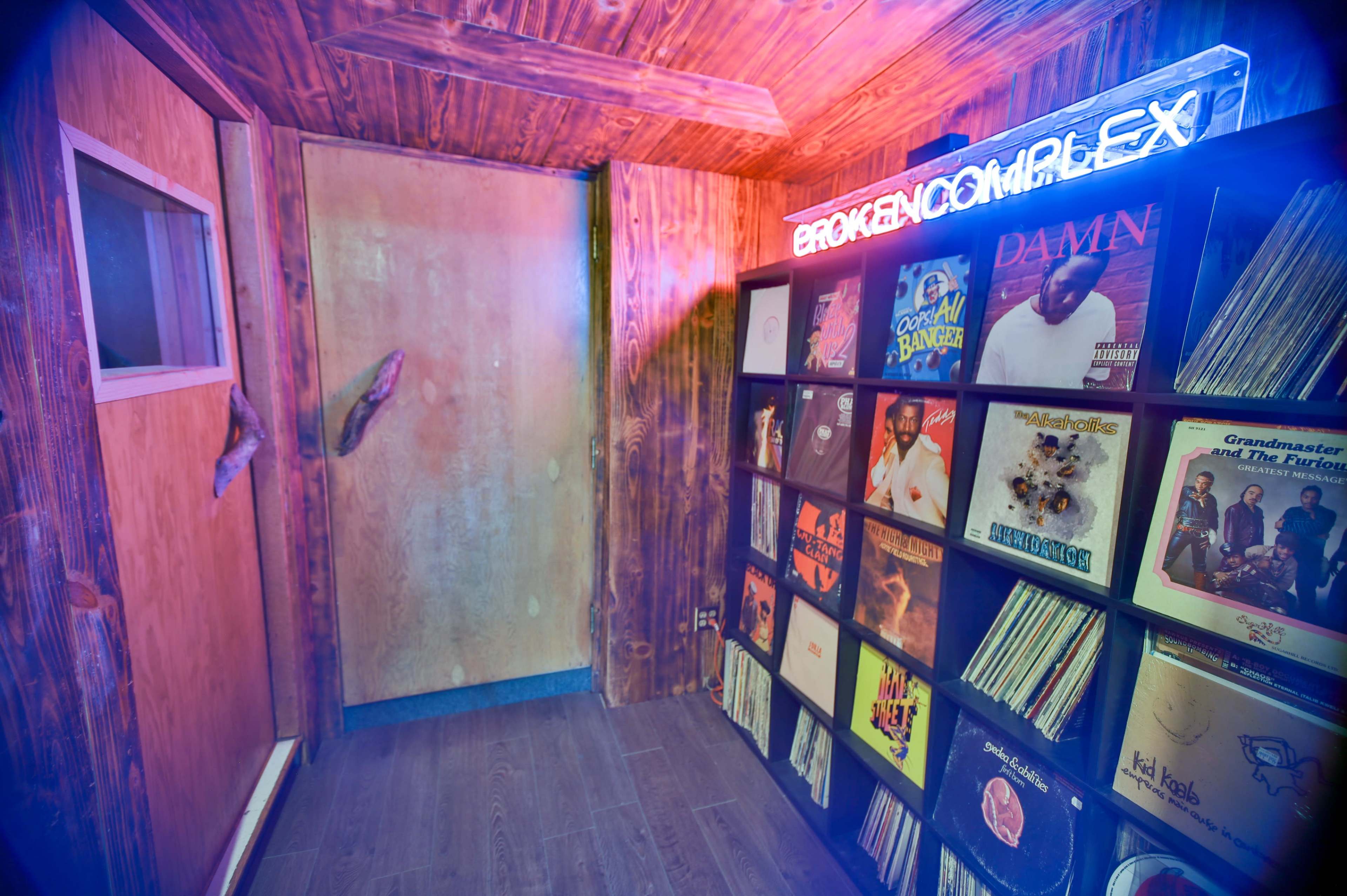 A small, wooden room featuring a wall of vinyl records and a neon sign that reads "BROKEN COMPLEX."