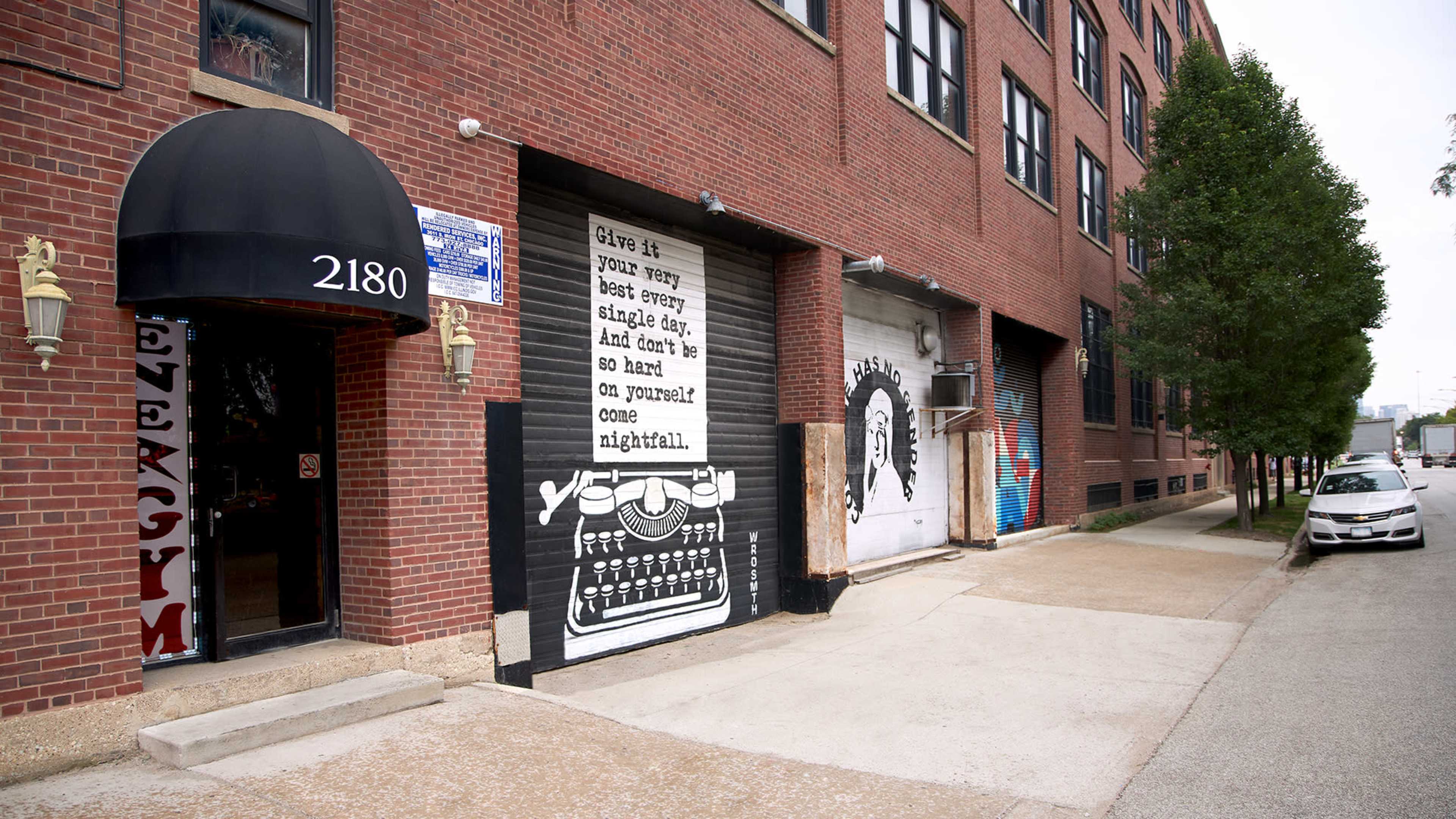 A brick building features a number of murals on its exterior, including a large painted typewriter and a quote on one wall, as well as signage for an establishment labeled "2180."