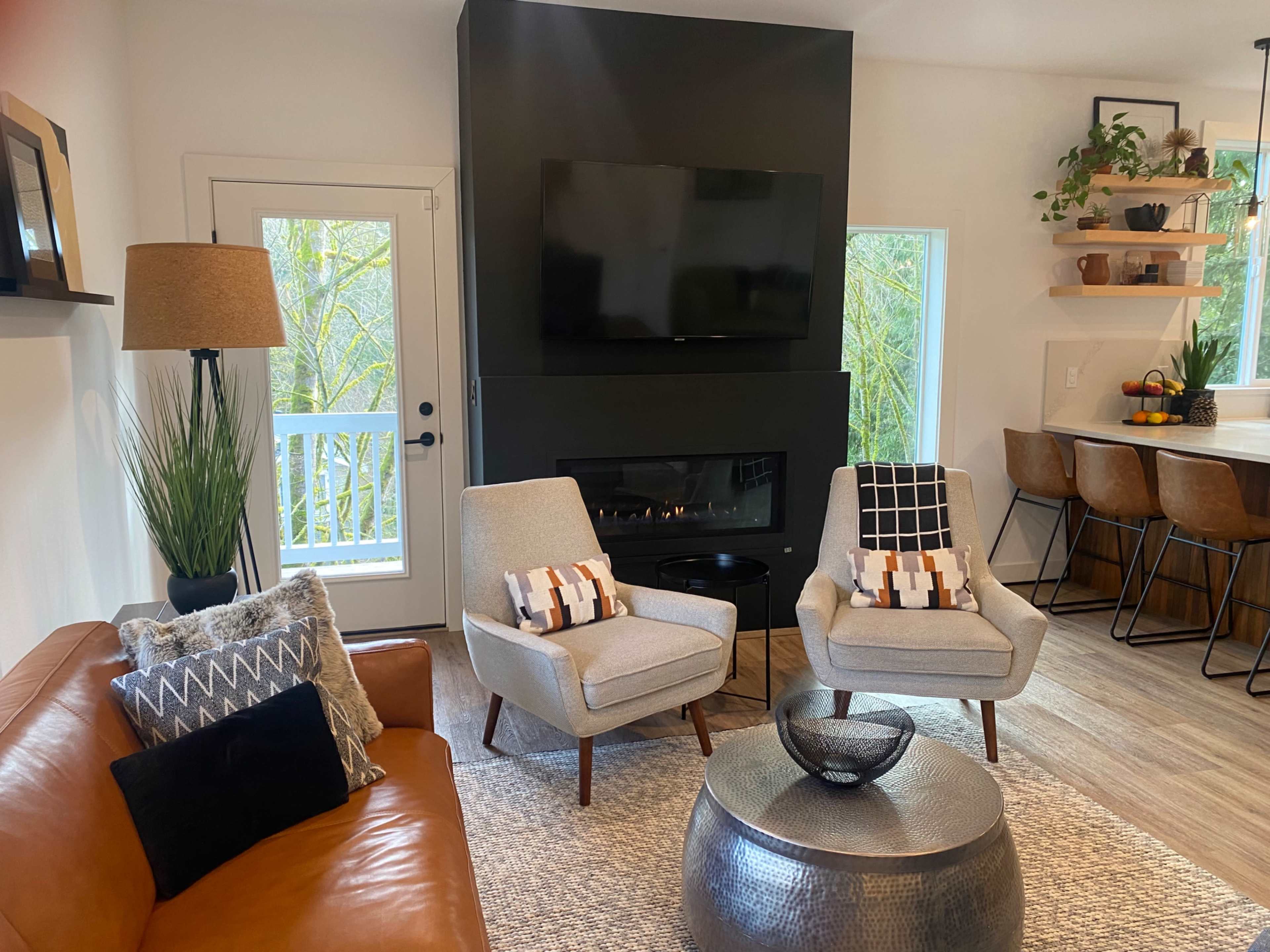 A modern living room with a brown leather sofa, two light-colored armchairs, a round metallic coffee table, a wall-mounted television, and a fireplace, all set against a backdrop of large windows.
