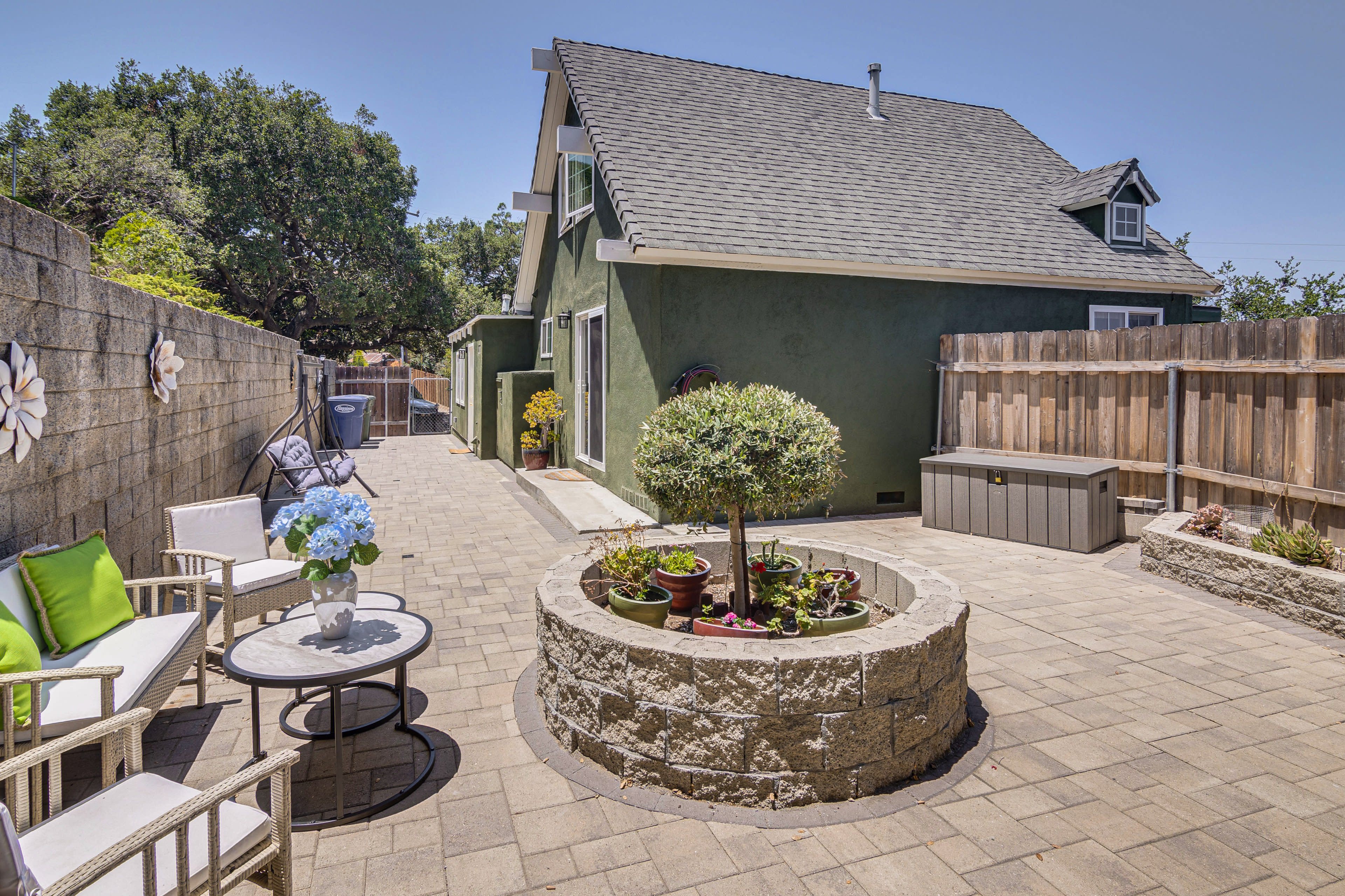 The image shows a backyard patio with a circular stone planter, outdoor seating, and a house with a green exterior.