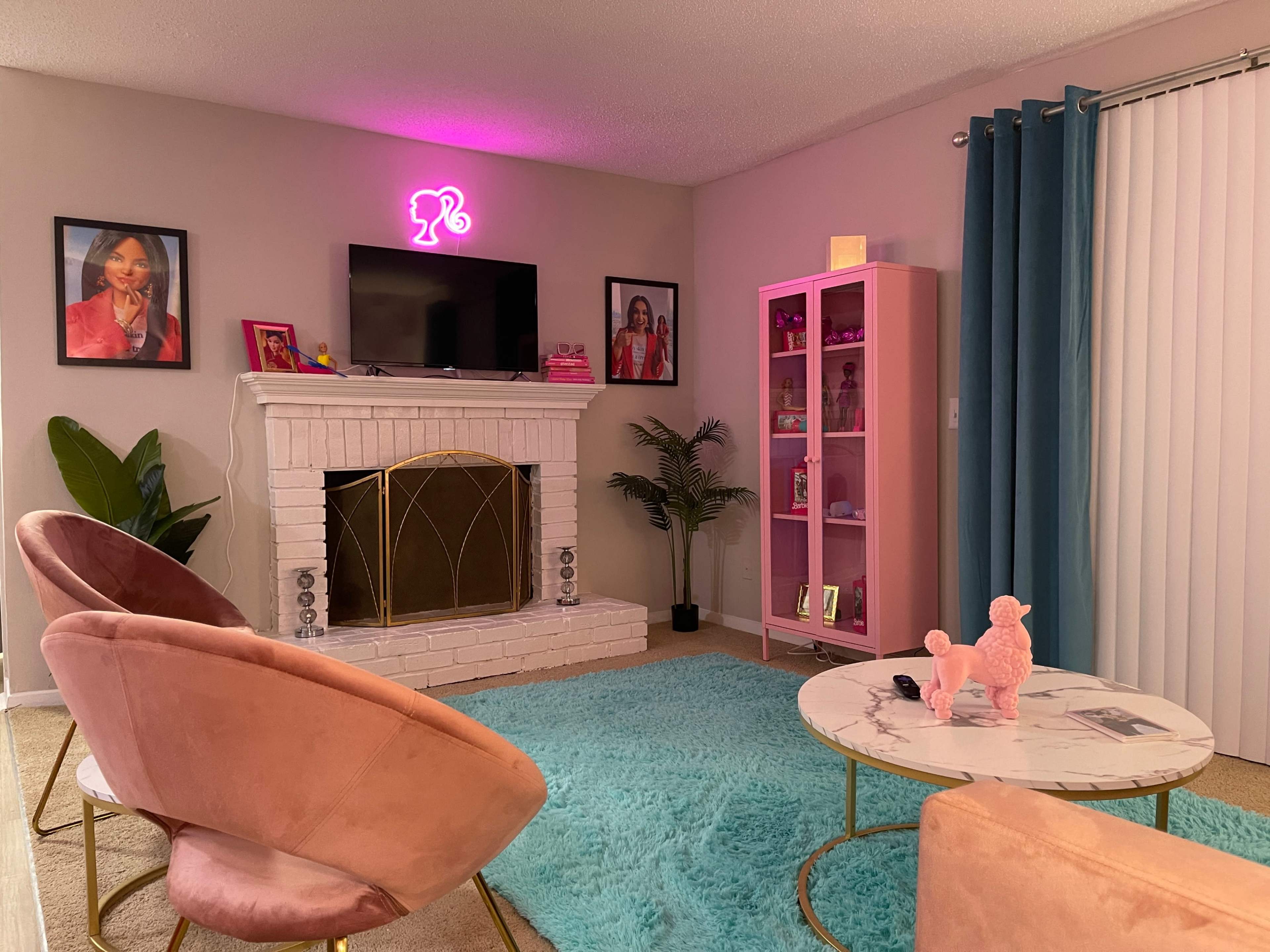 A cozy living room features a pink neon sign, framed pictures on the wall, a white brick fireplace, and modern furniture including a round coffee table and plush armchairs.
