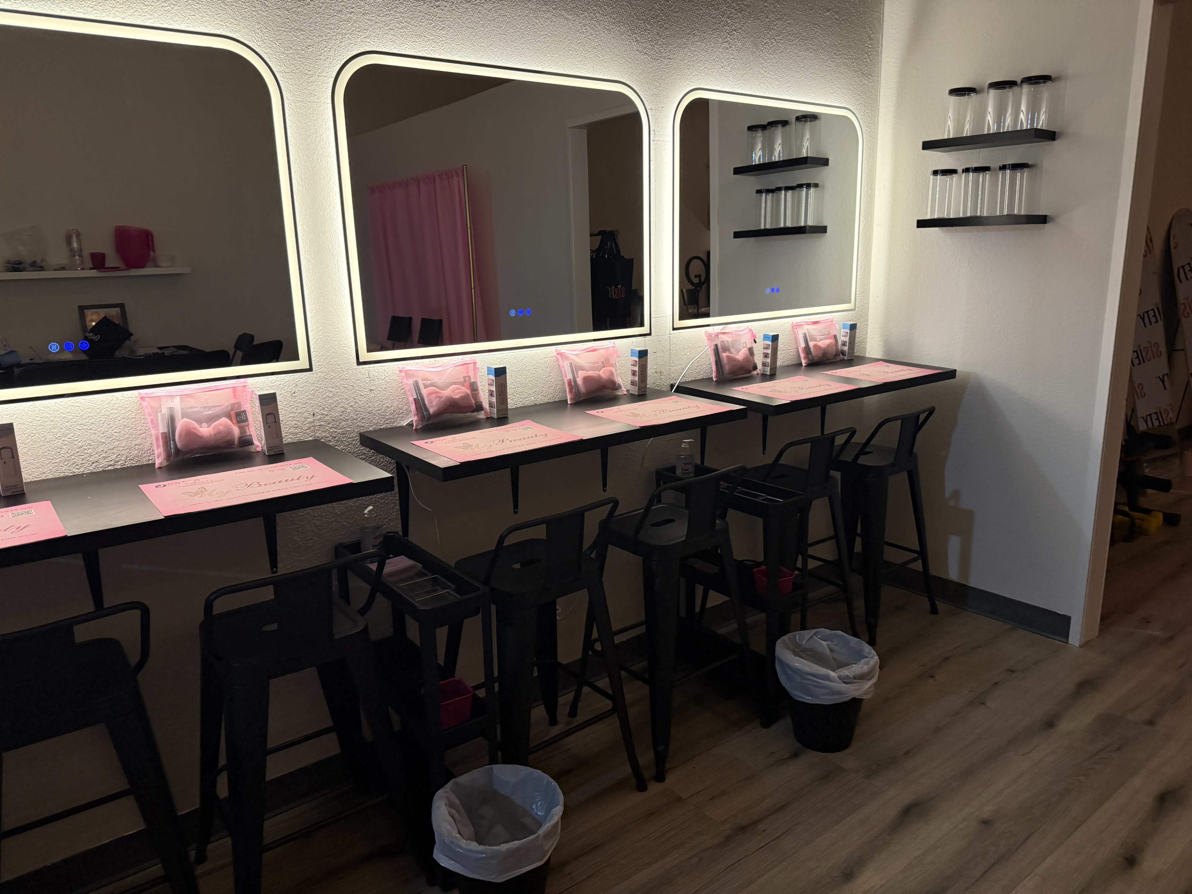 The image shows a salon or beauty studio with three mirrored stations, each equipped with a black chair, a pink table setting, and a waste bin underneath.