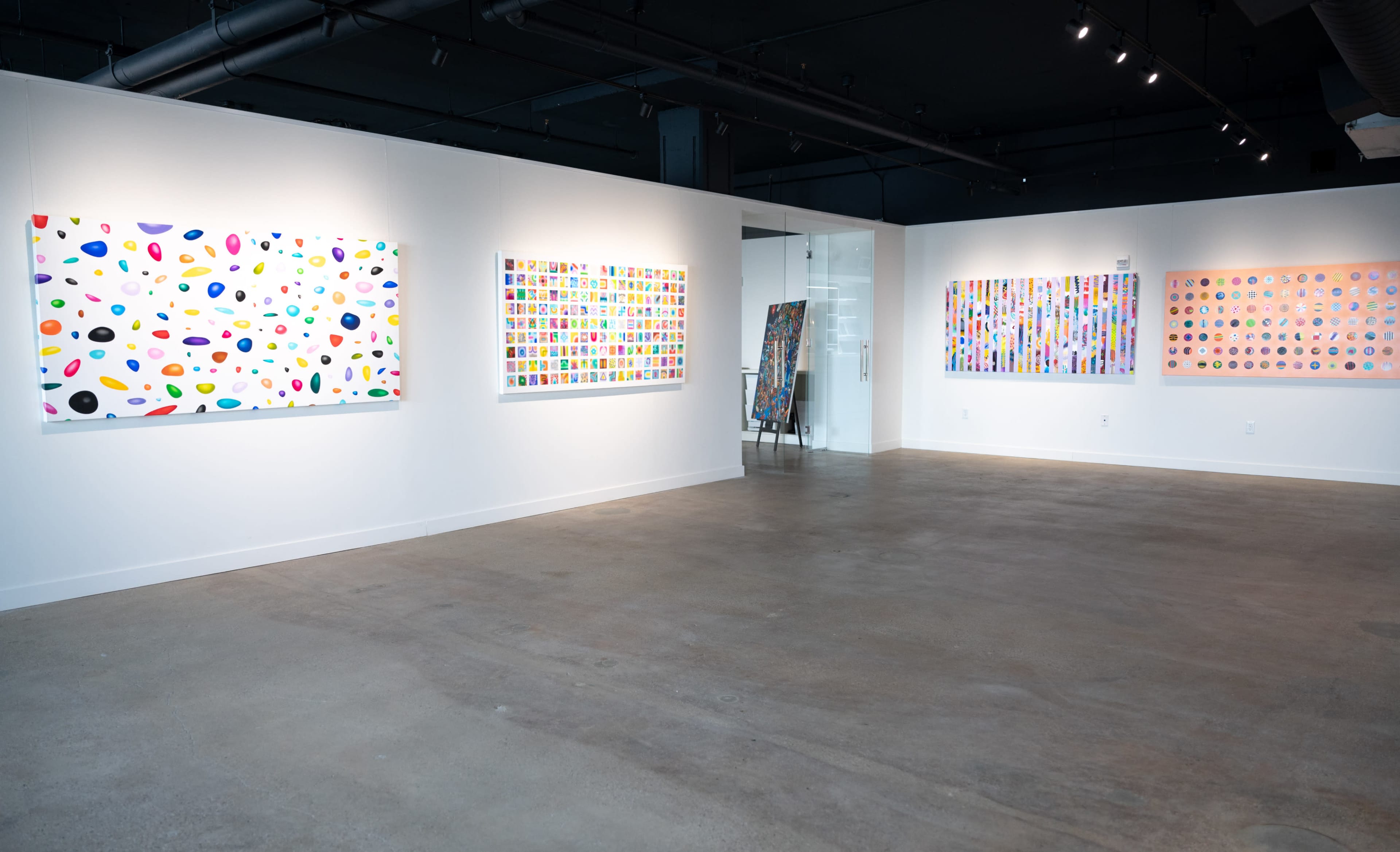An art gallery features three vibrant abstract paintings displayed on white walls, with a spacious concrete floor and modern lighting.