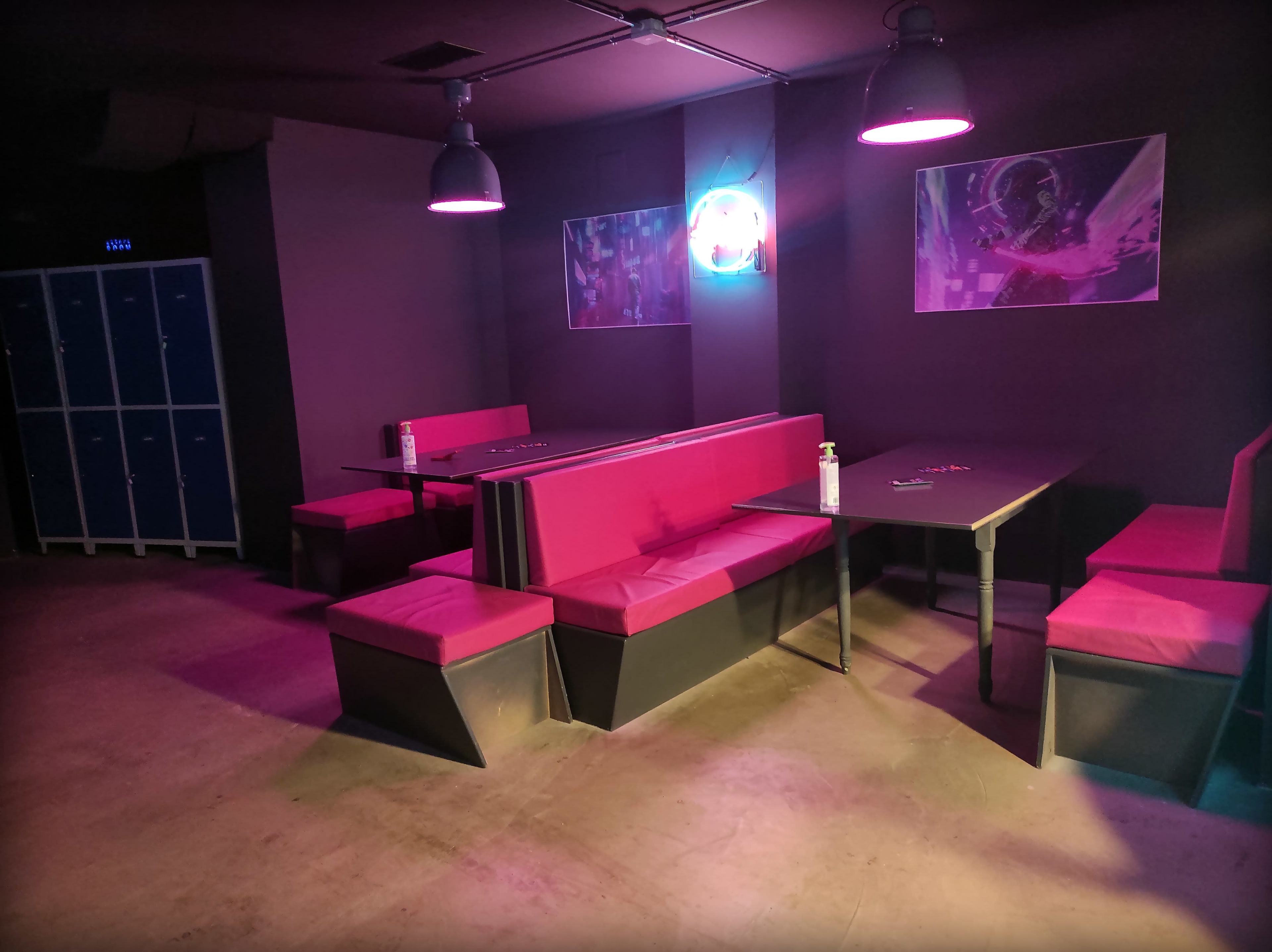 A dimly lit room with pink seating and a table, accompanied by neon lights and lockers in the background.