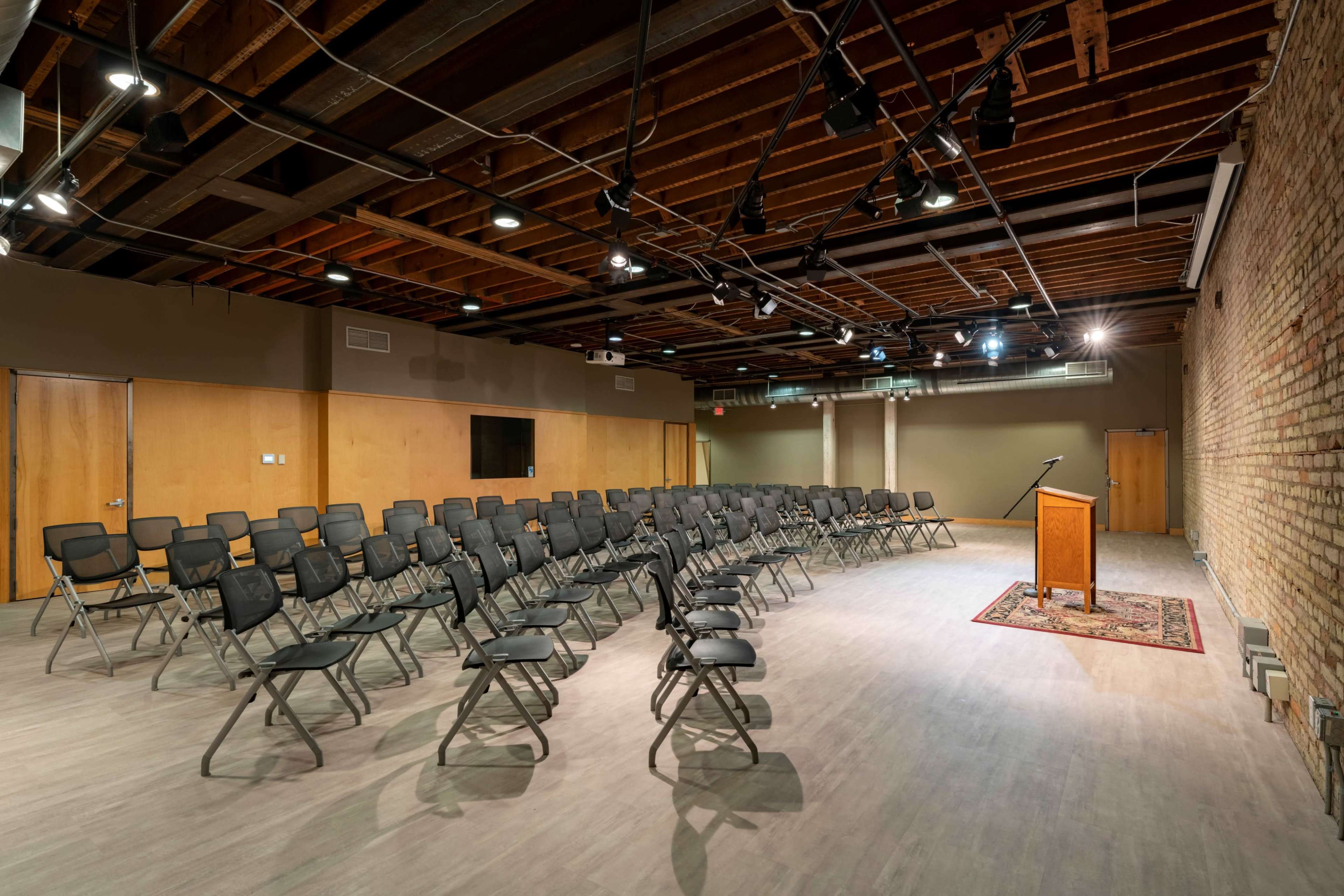 The image shows a spacious room with rows of black folding chairs arranged facing a wooden podium on a rug, with exposed brick walls and overhead lighting.
