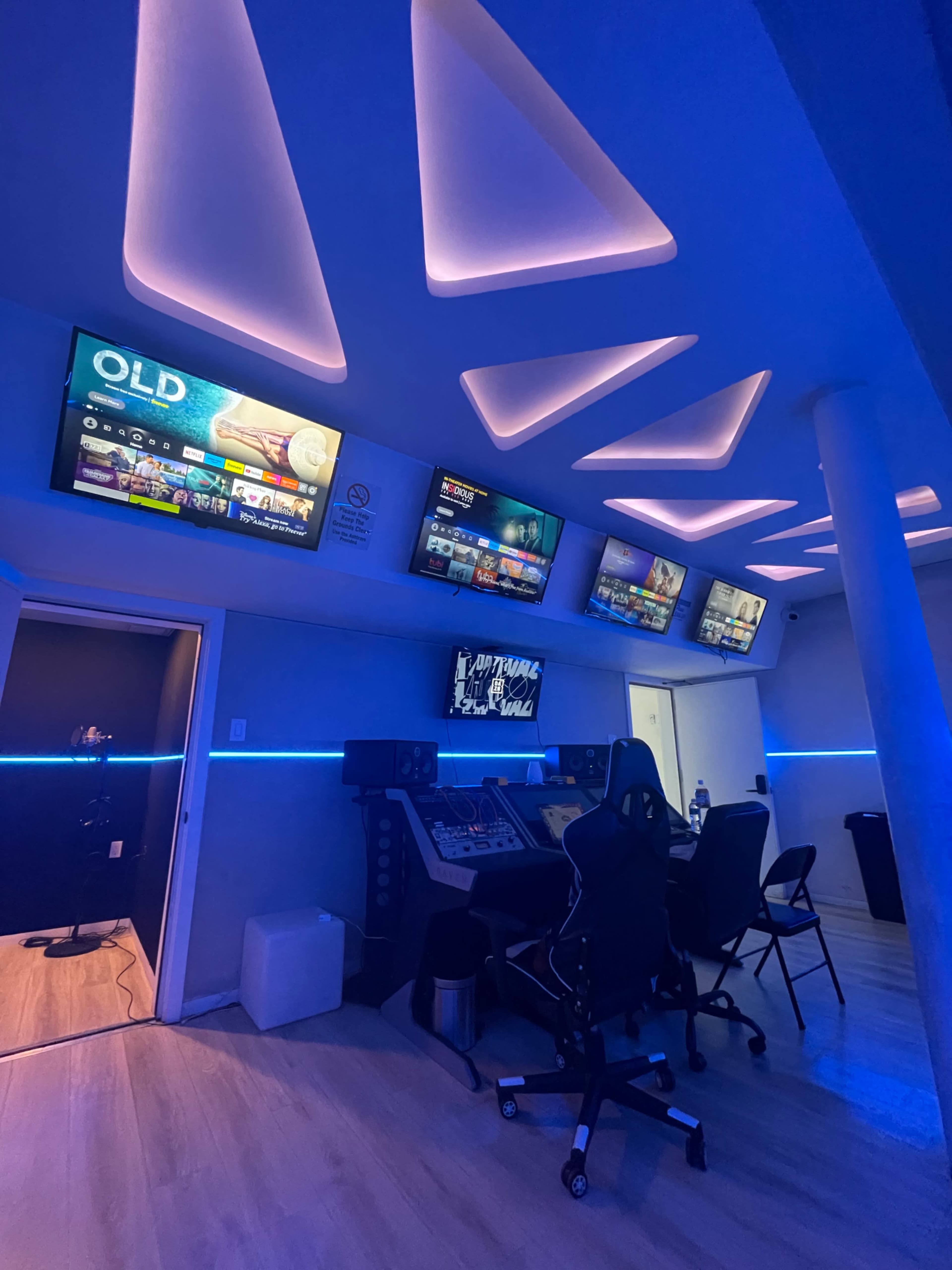 A modern gaming or multimedia room featuring multiple screens mounted on the wall, ambient blue lighting, and a setup with chairs and equipment.