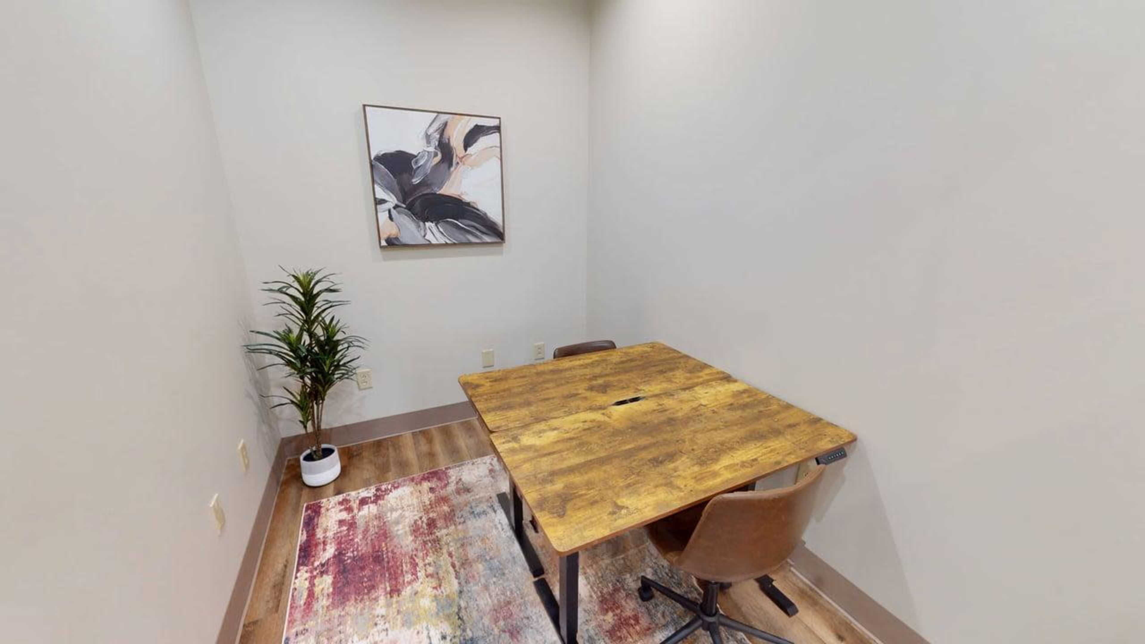 A small room features a wooden table and two chairs, with a piece of abstract art on the wall and a potted plant in the corner.