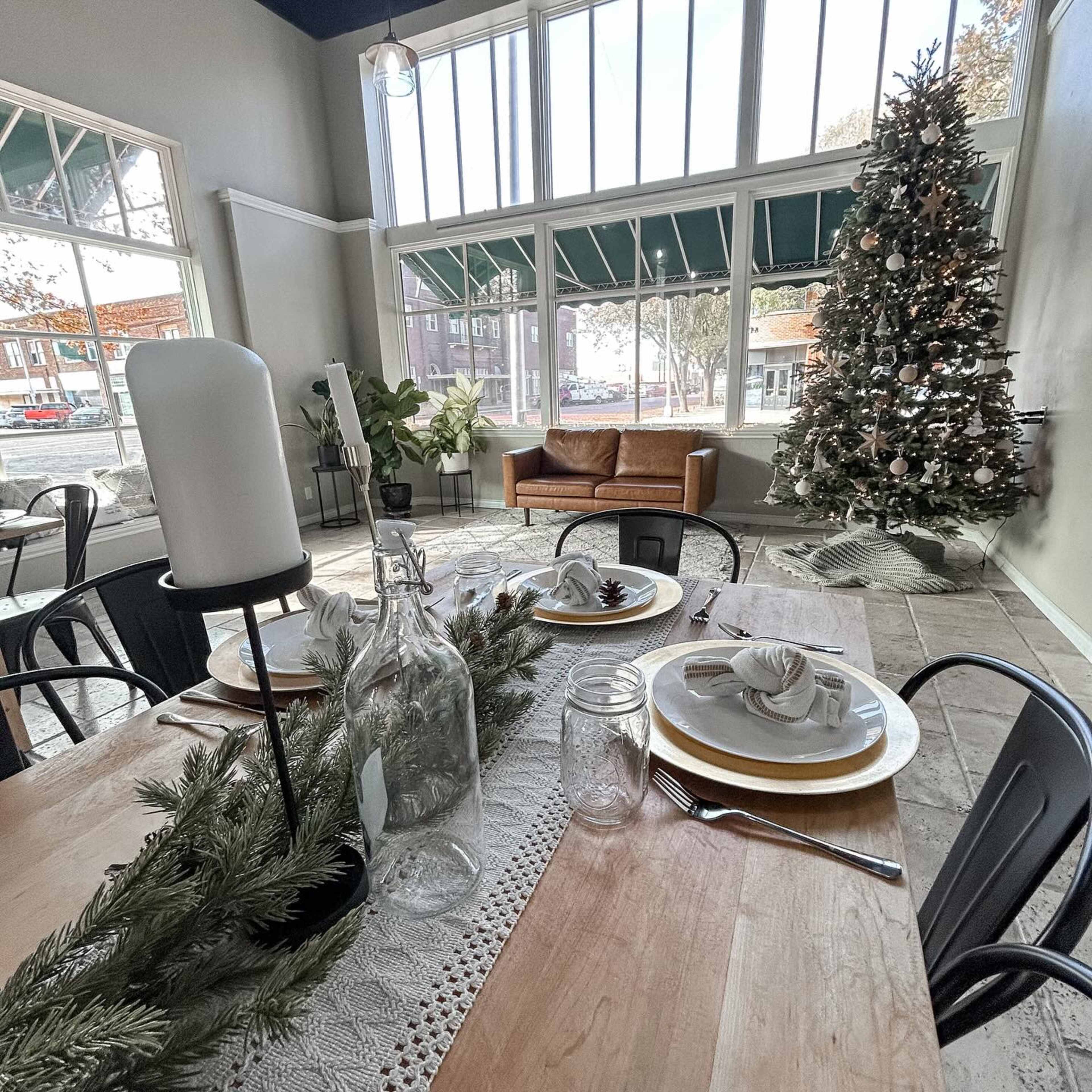 A dining table set with plates and napkins in a room featuring large windows, a decorated Christmas tree, and a cozy seating area.