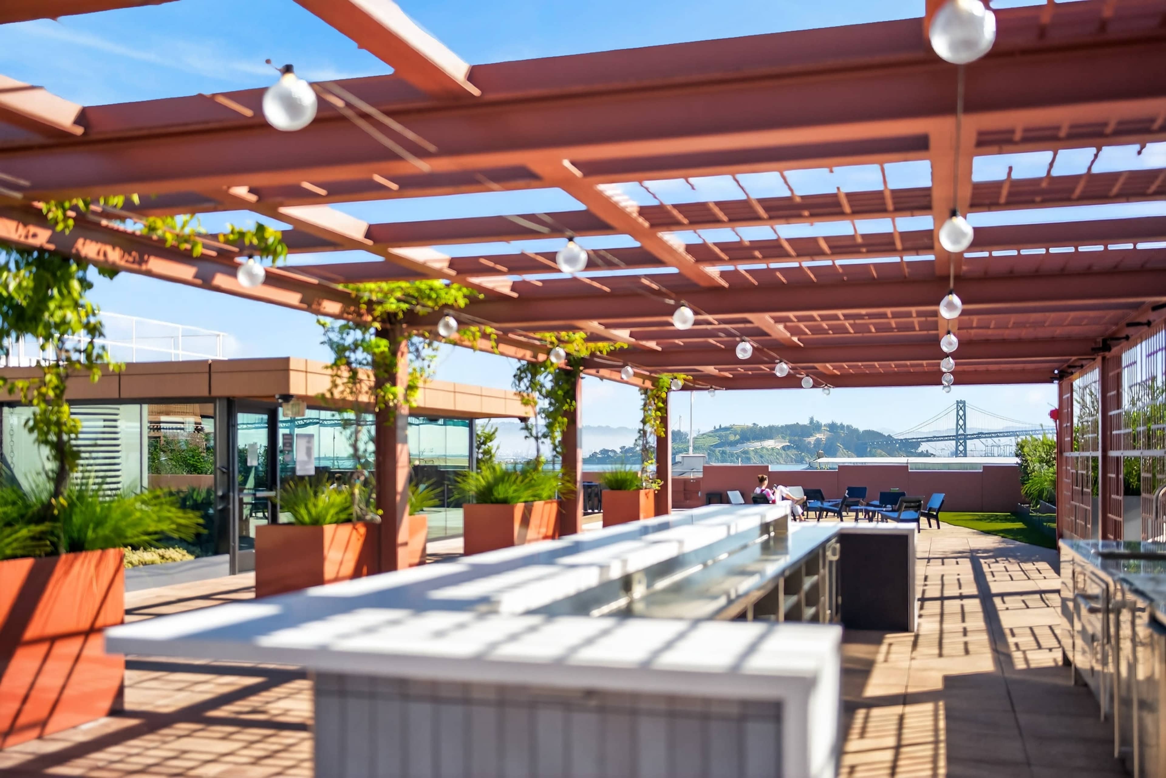 The image shows a rooftop terrace with wooden pergolas, hanging lights, and planters, overlooking a bridge and distant hills.