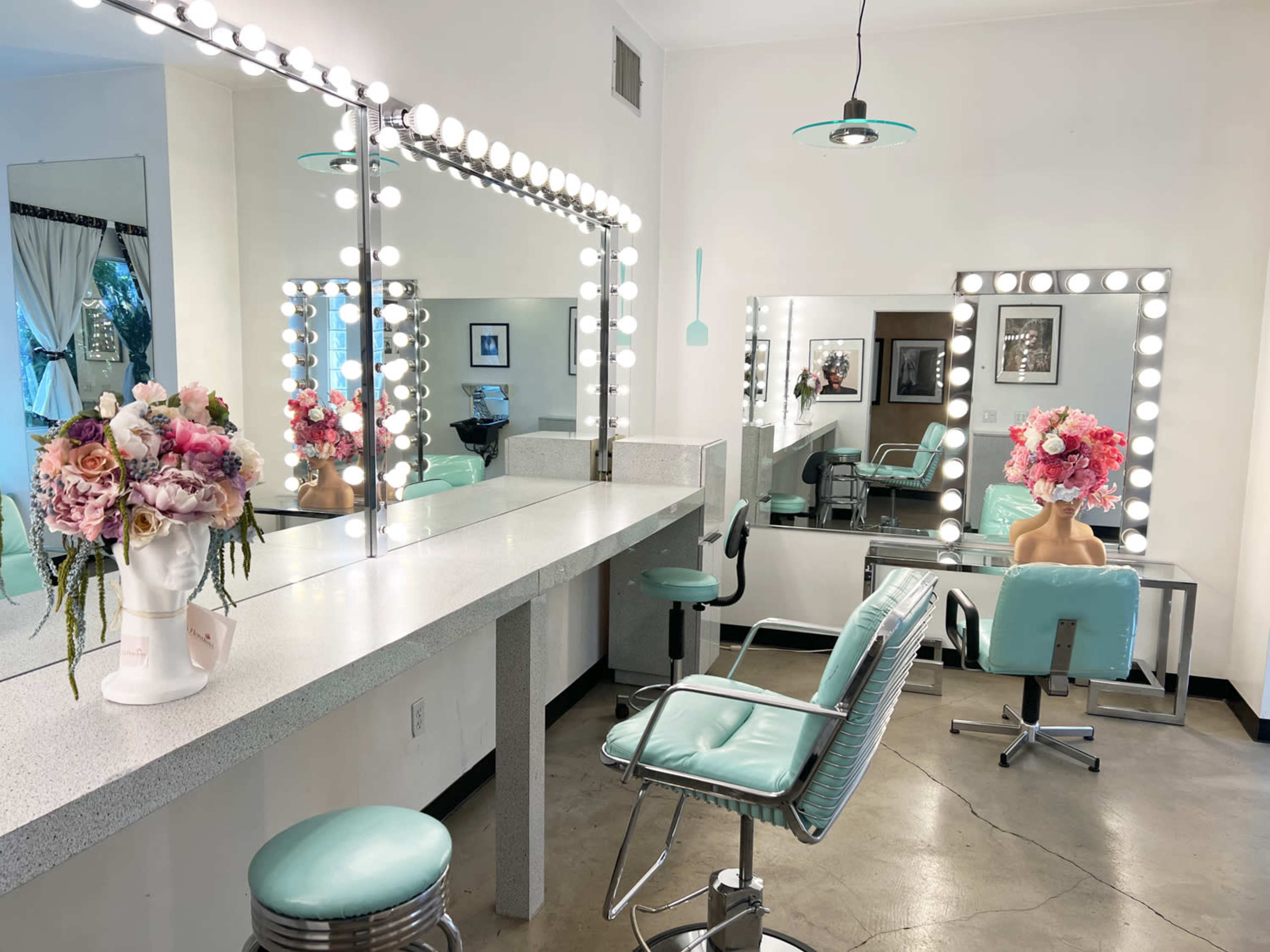 The image shows a brightly lit hair salon with a large mirror, a marble countertop, turquoise chairs, and floral arrangements placed on the counters.