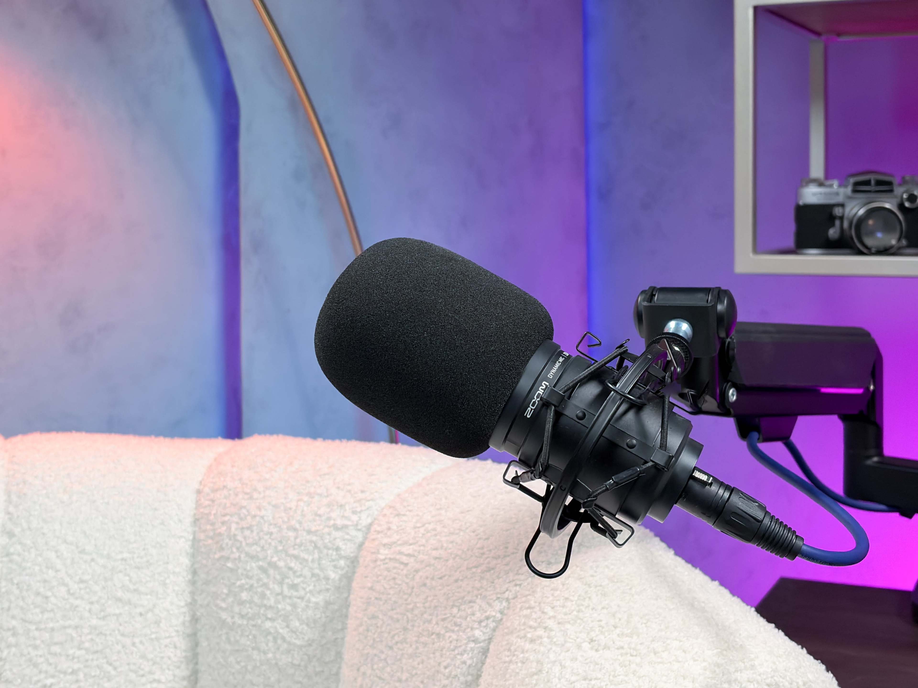A black microphone is mounted on a stand next to a white, textured couch against a colorful background.