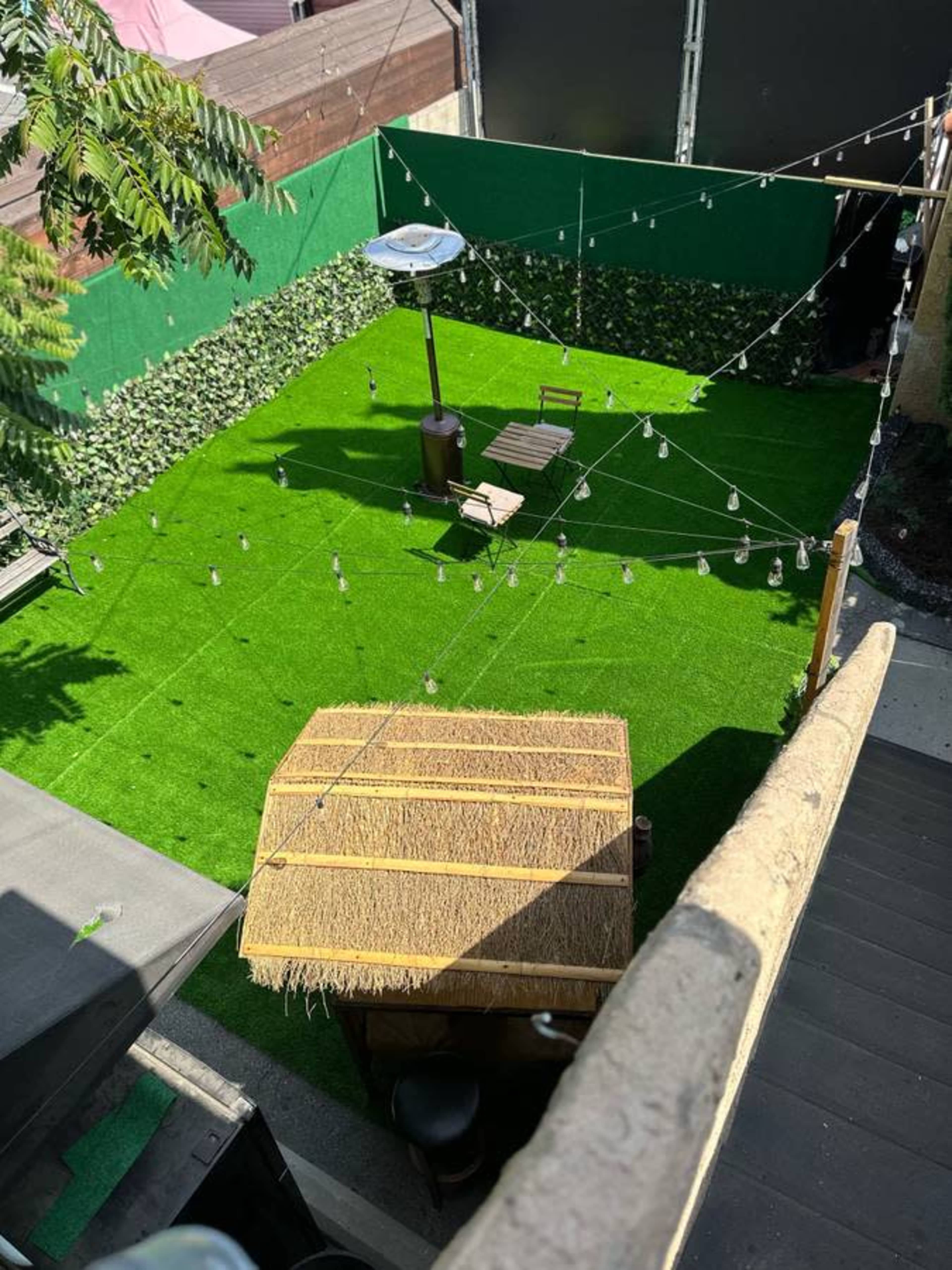 A green, artificial turf backyard featuring a thatched-roof structure, two wooden chairs, and string lights overhead.