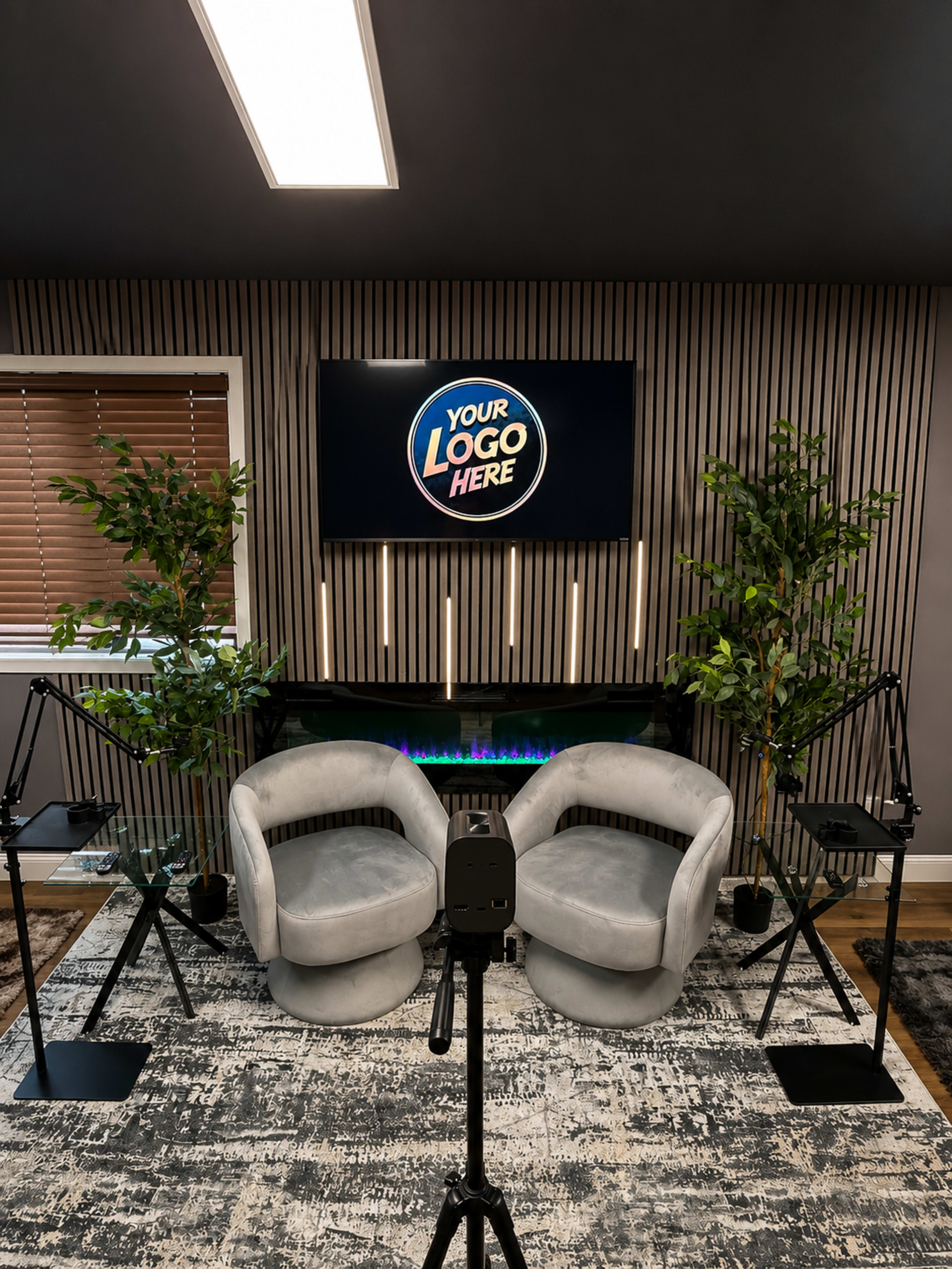 The image shows a modern studio setup with two gray chairs facing a large screen displaying the text "YOUR LOGO HERE," flanked by two potted plants and a decorative electric fireplace.