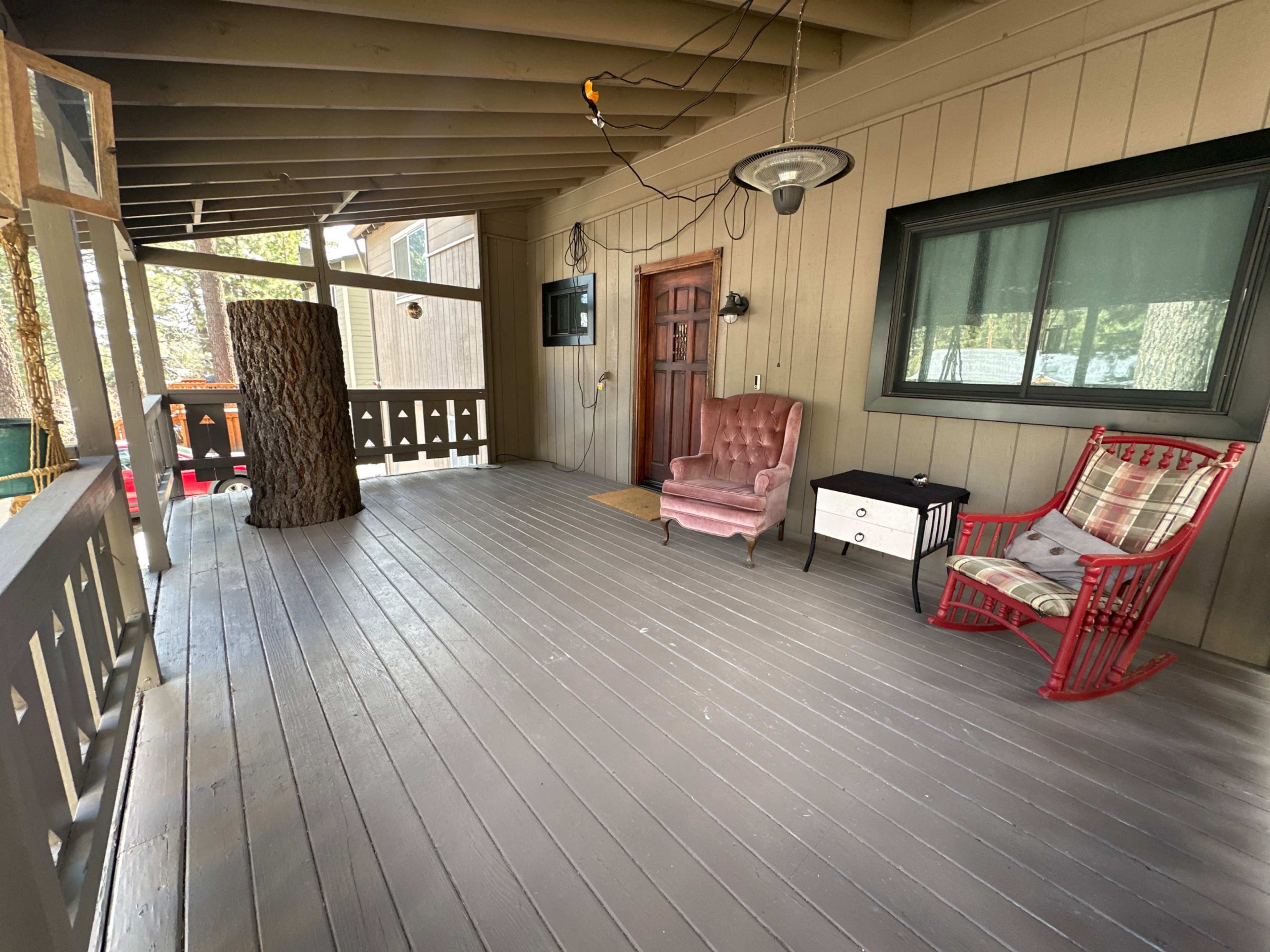A covered porch with wooden flooring, featuring a rustic tree trunk, a pink armchair, a small black and white table, and a red rocking chair against a backdrop of wooden walls and a window.