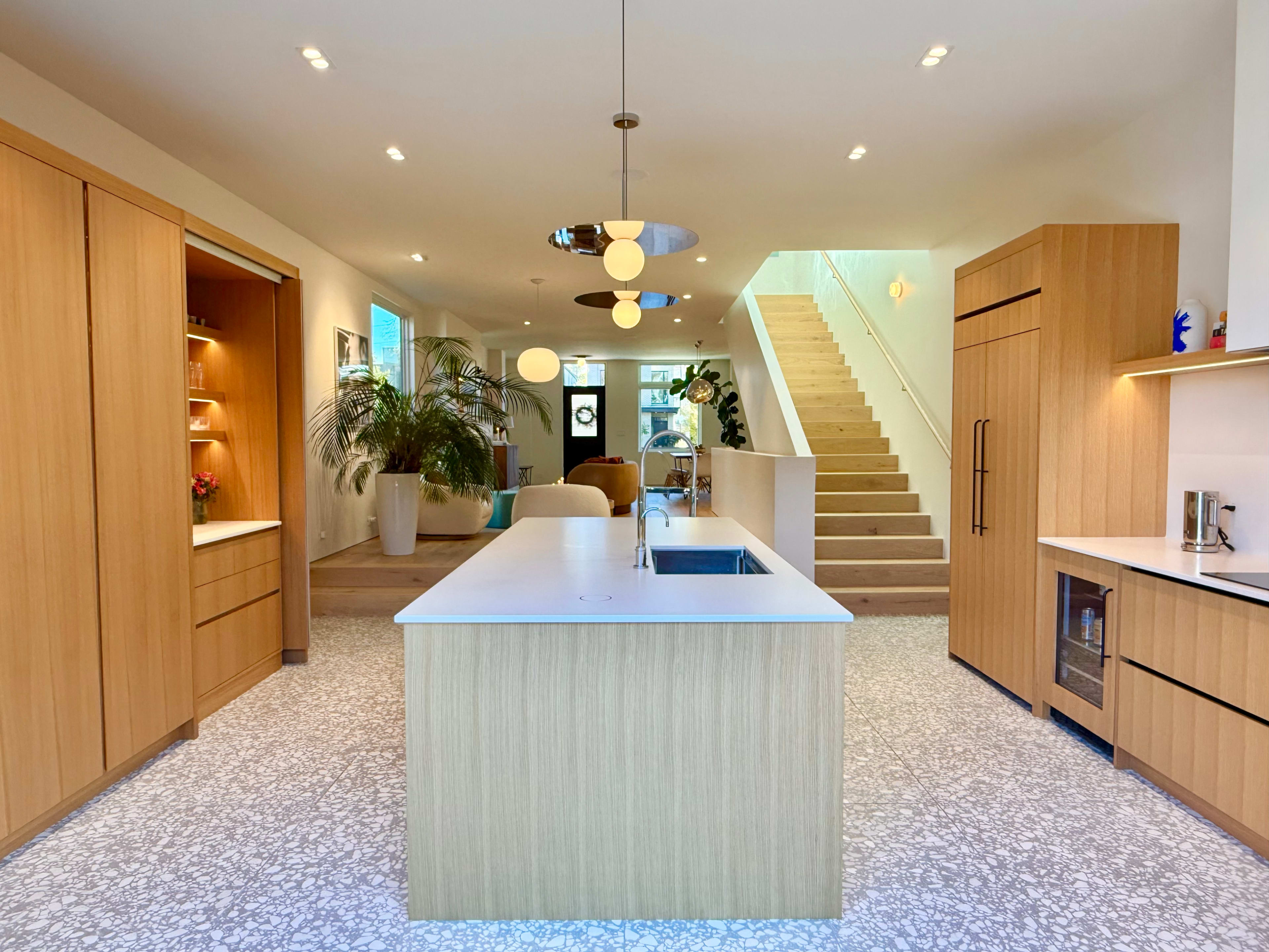 The image shows a modern kitchen with a central island, wooden cabinetry, a stairway in the background, and large windows allowing natural light to enter.