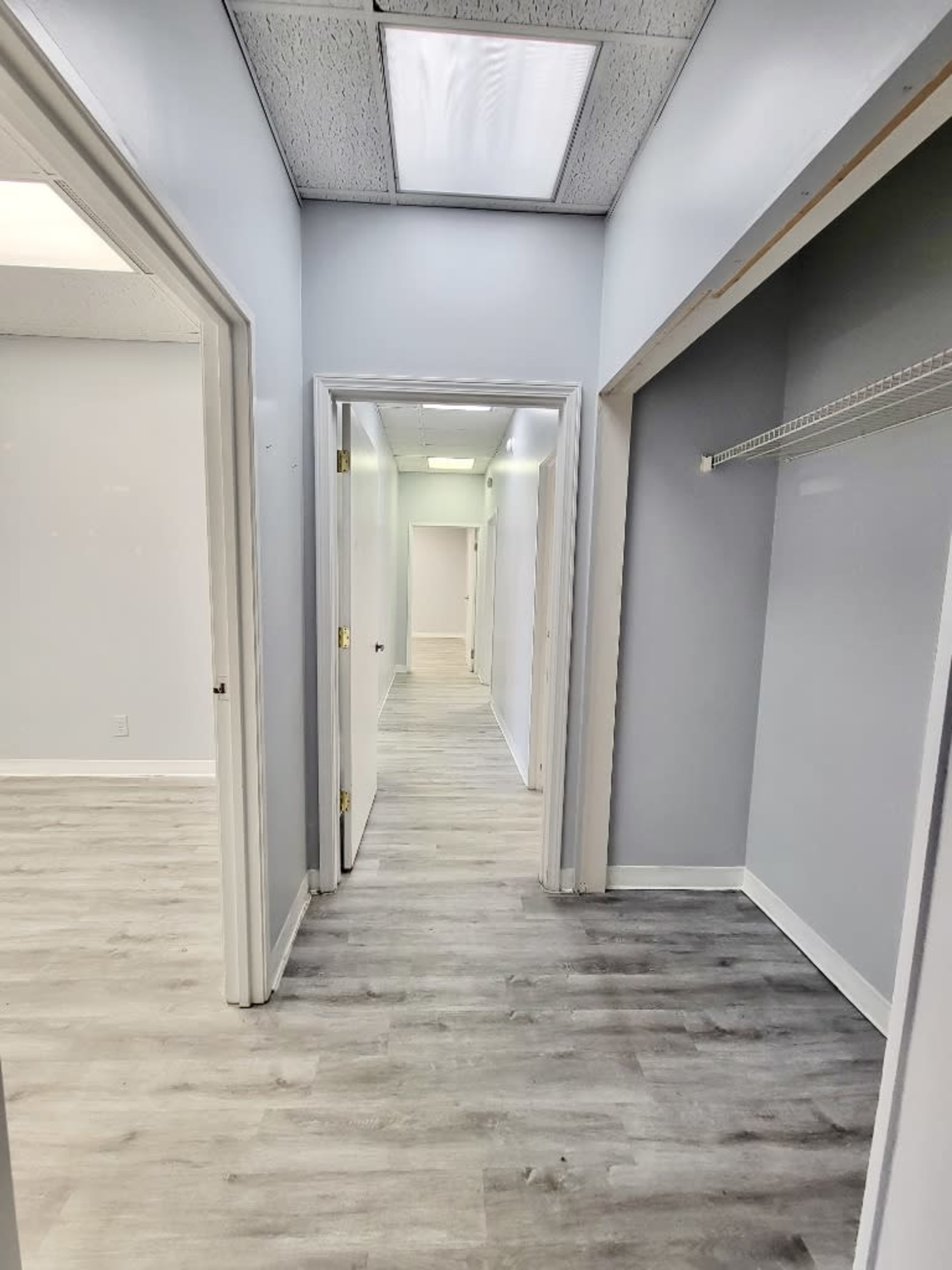 A long hallway with gray walls and light wooden flooring, leading to doors on either side and a bright area at the end.