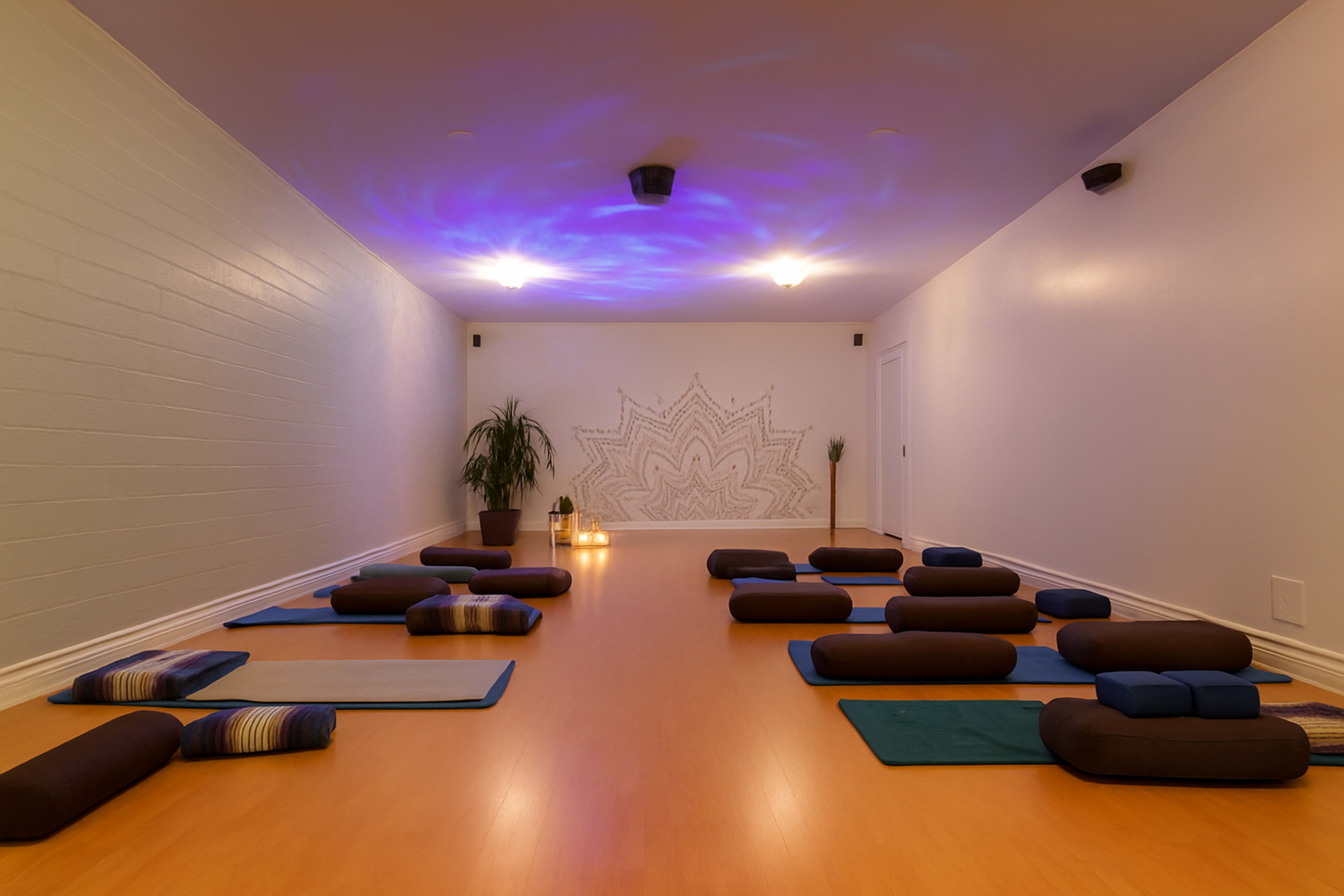The image shows a serene yoga studio with mats and bolsters arranged neatly on a wooden floor, illuminated by soft lighting.