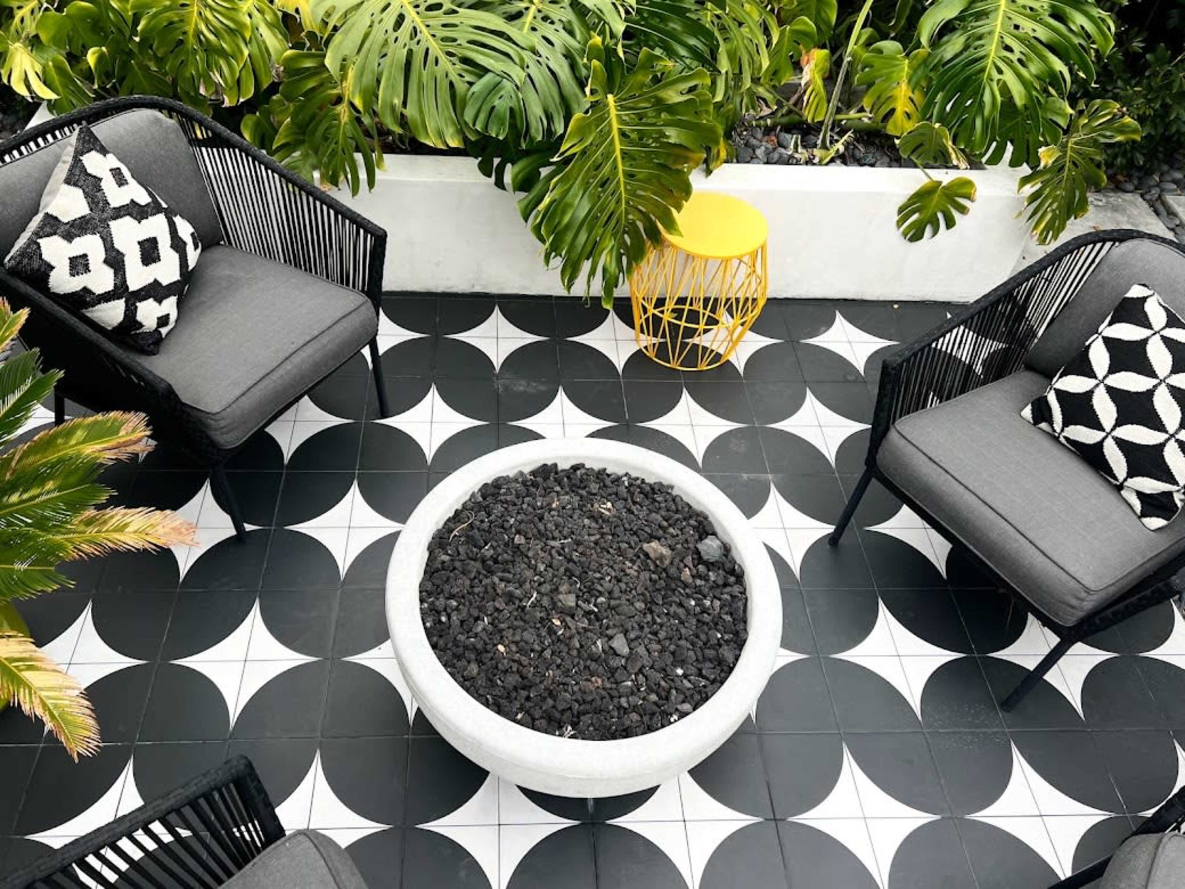 The image shows a cozy outdoor seating area featuring two black chairs with patterned cushions, a round concrete fire pit filled with black stones, and a yellow side table, all set on a geometric black and white tiled floor surrounded by tropical plants.