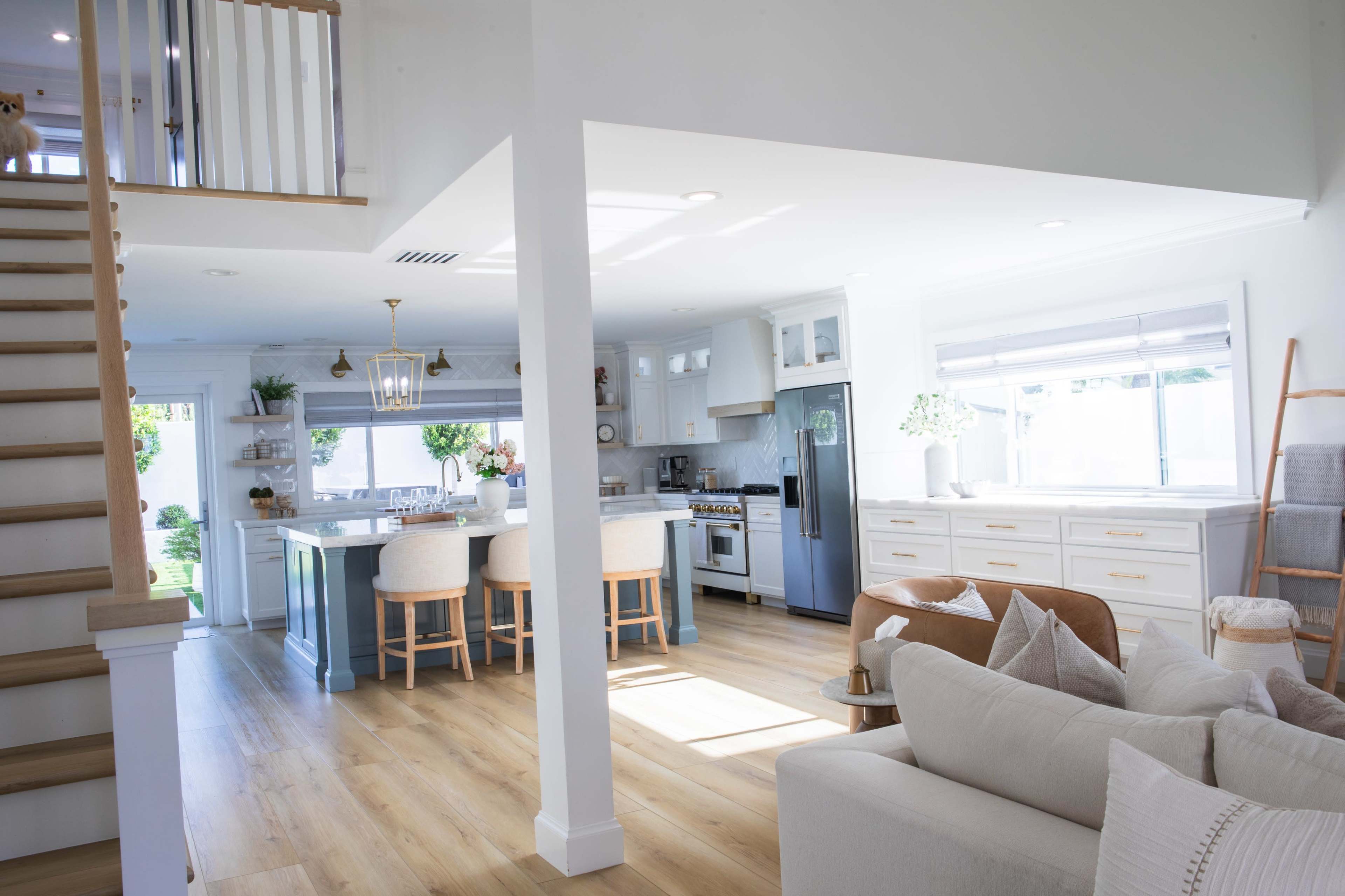 The image shows a bright, modern kitchen and living area with wooden flooring, white cabinetry, and a staircase leading to a second floor.