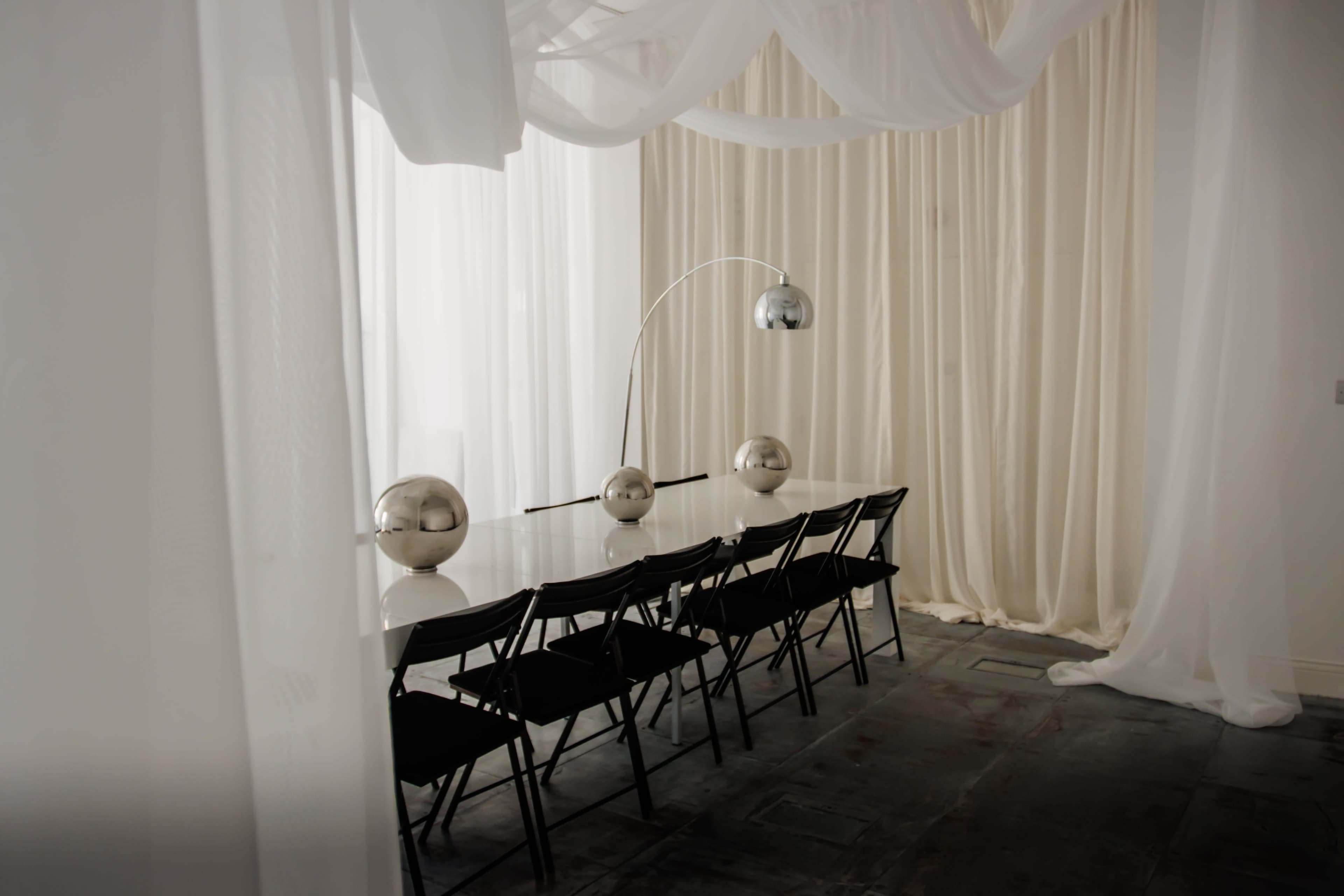 A set of black chairs lines a long white table under a curved metallic lamp in a room with sheer white drapery.
