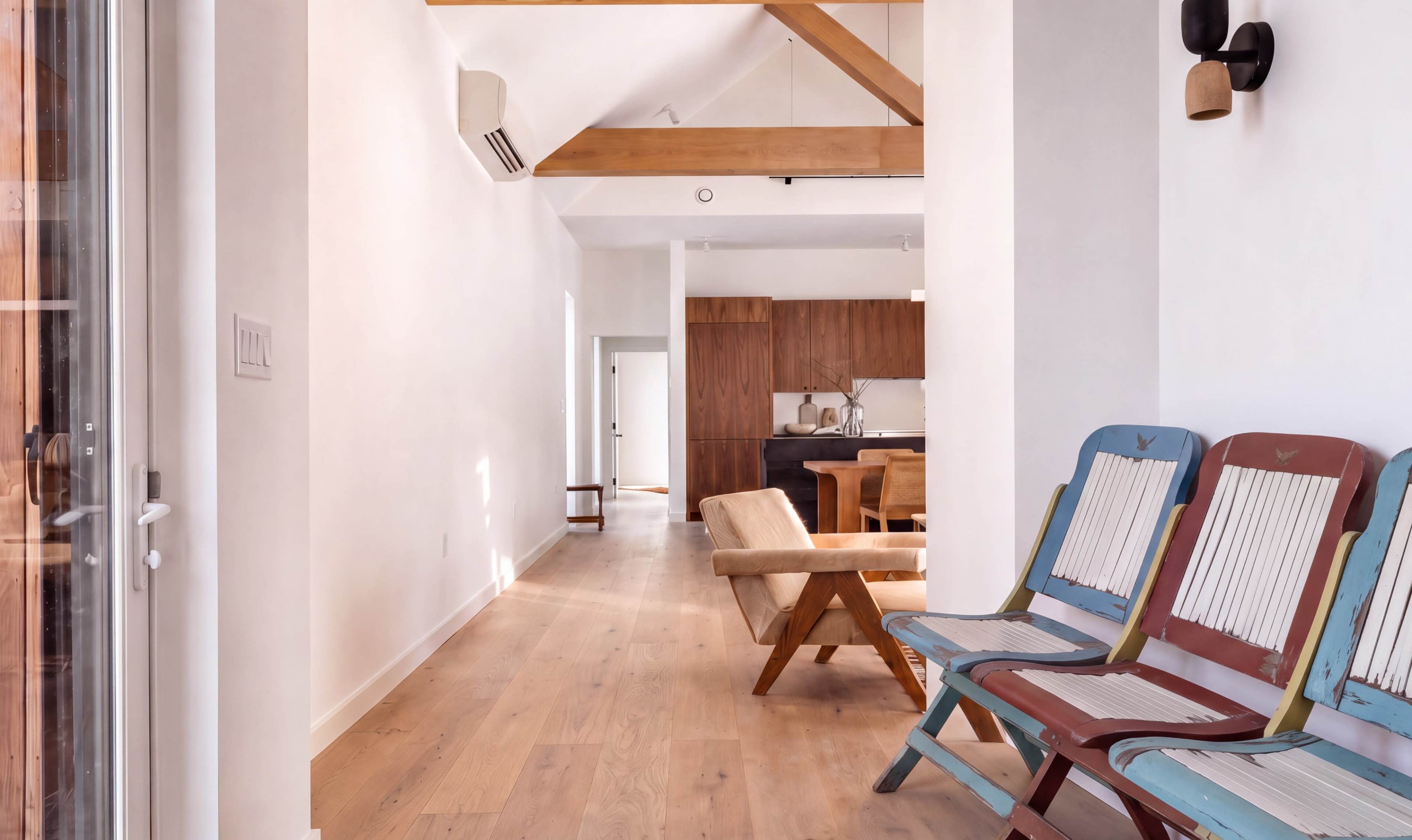 The image features a bright hallway with wooden beams, leading to a modern kitchen, and includes colorful, vintage-style chairs aligned against one wall.
