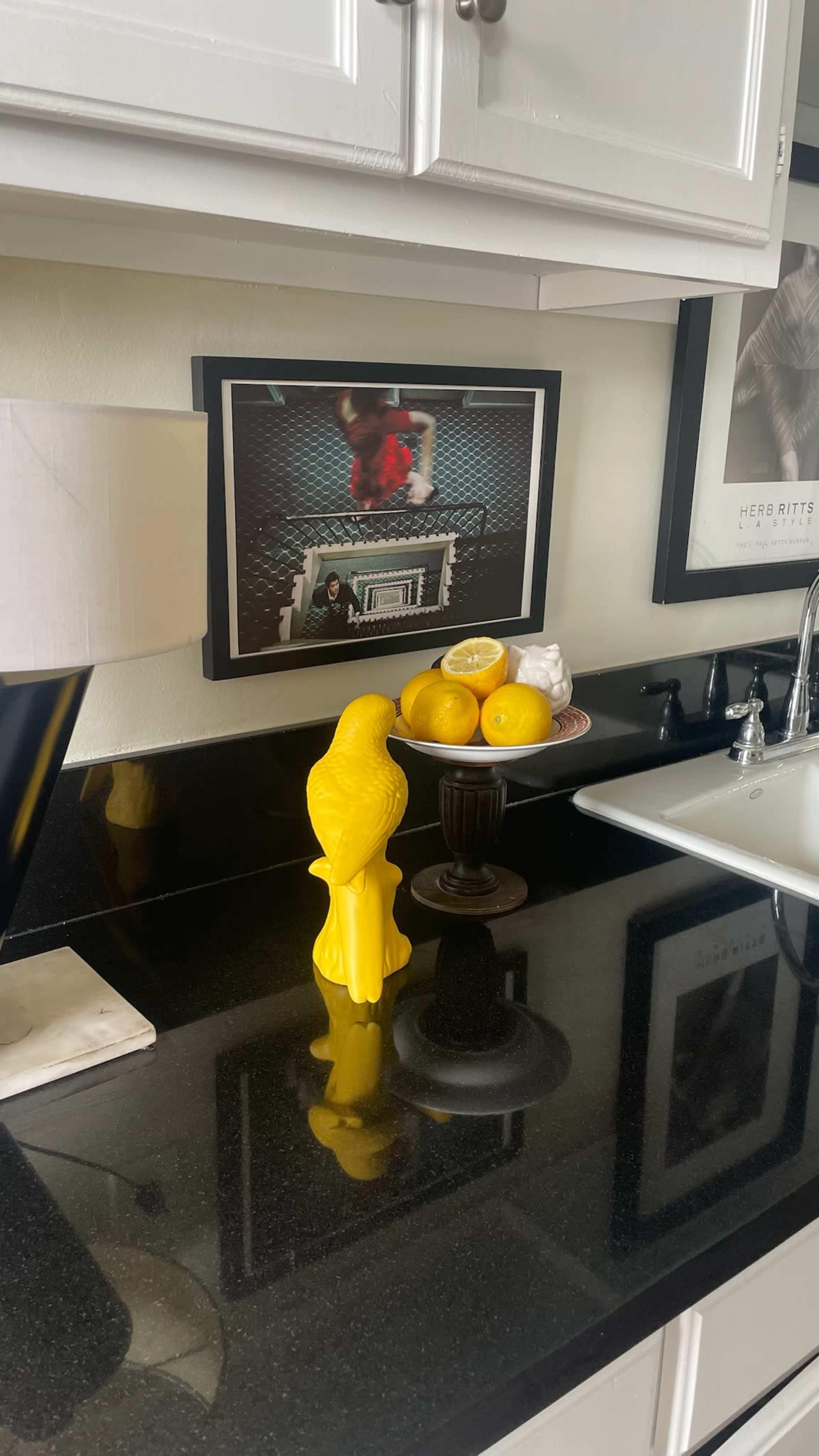 A yellow decorative bird and a bowl of lemons sit on a black countertop next to a framed photograph and a lamp.