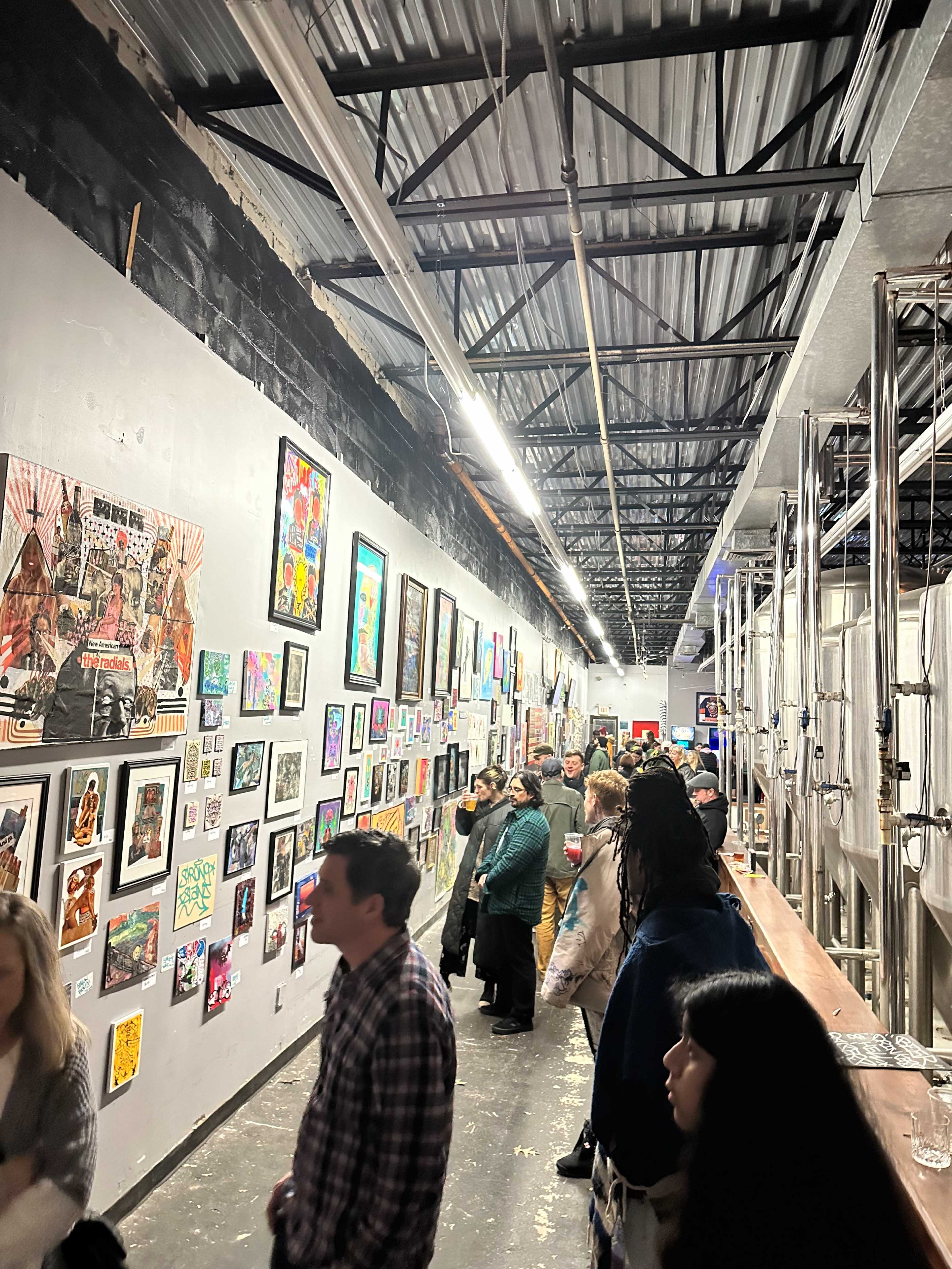 A crowded art gallery showcases a variety of framed artworks on the walls, with attendees interacting and observing the pieces.