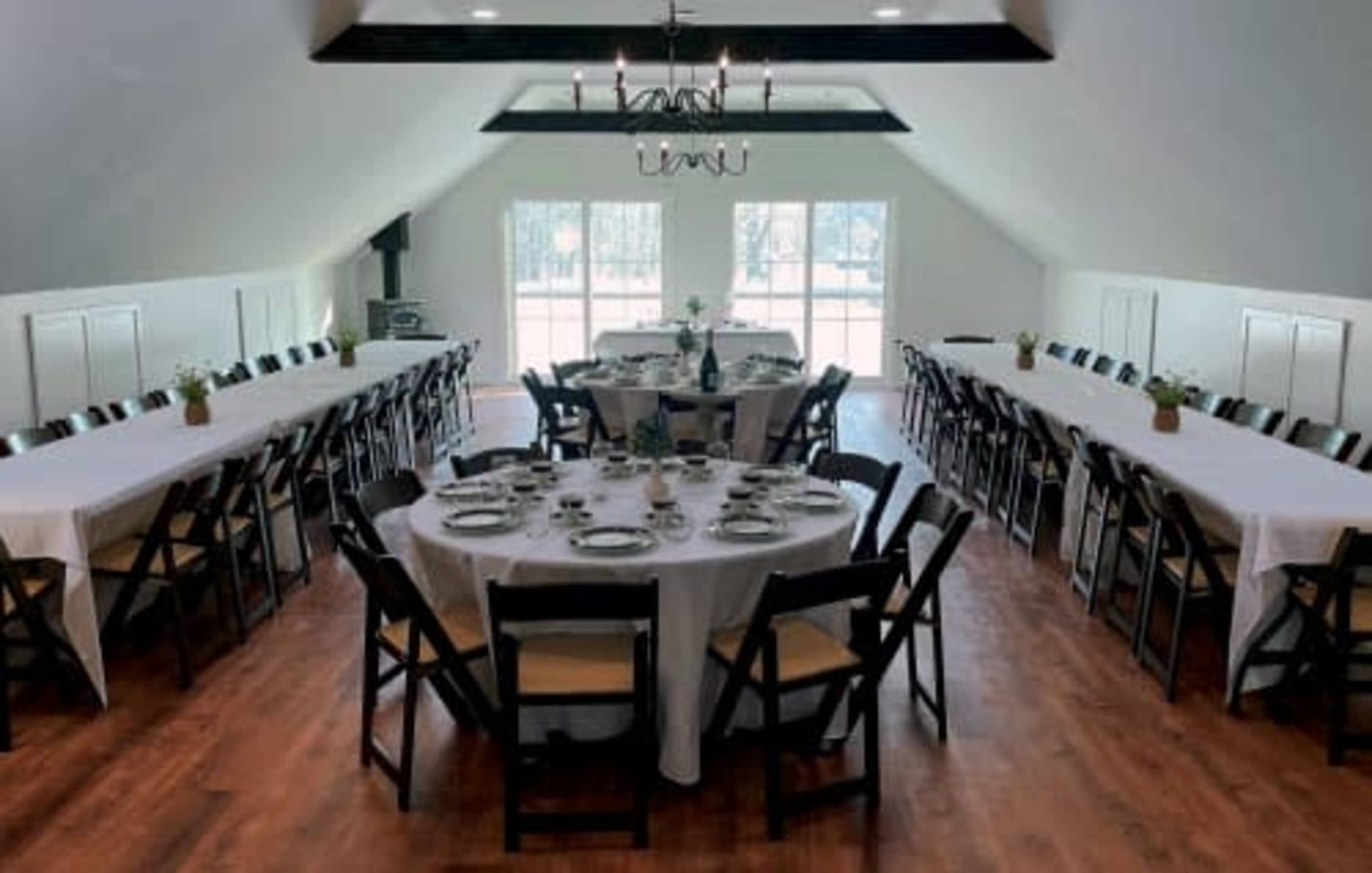 A spacious dining area features long tables set for a meal, with a chandelier hanging from the ceiling and large windows providing natural light.