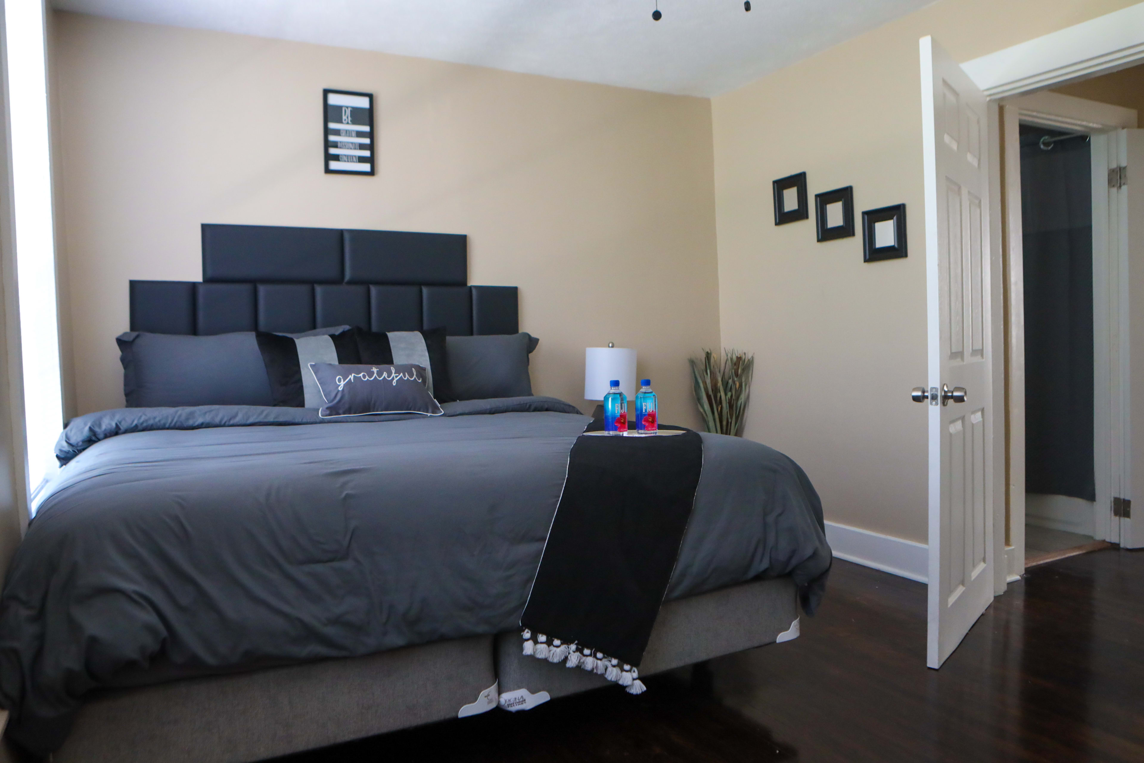 A modern bedroom features a large bed with a dark gray comforter, pillows, and a decorative throw, complemented by a bedside table with water bottles and a door leading to a bathroom.
