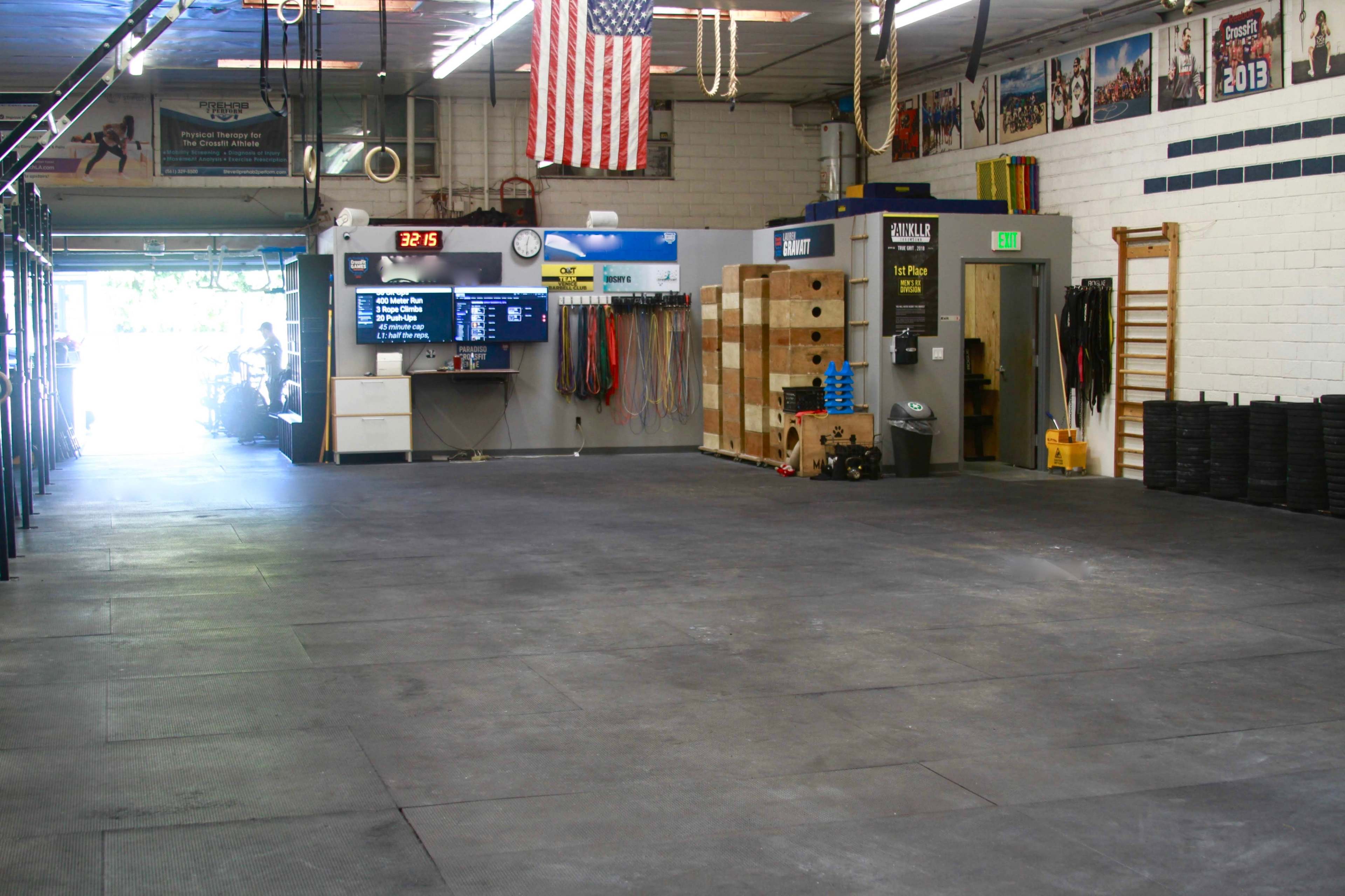 The image shows a spacious indoor gym with rubber flooring, exercise equipment, and a large display monitor on the wall.