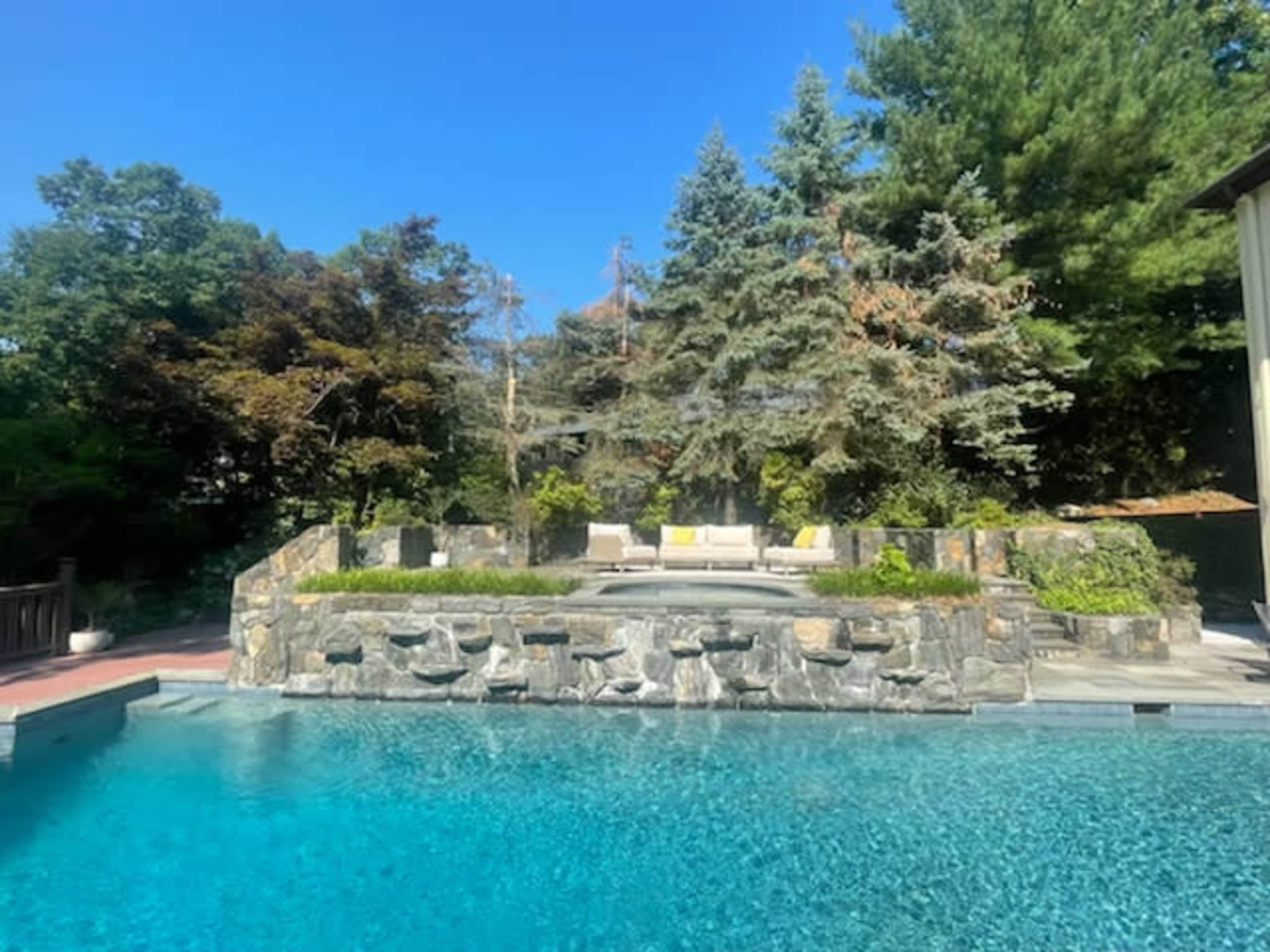 The image shows a pool surrounded by a stone wall and landscaped gardens, with a seating area visible in the background.