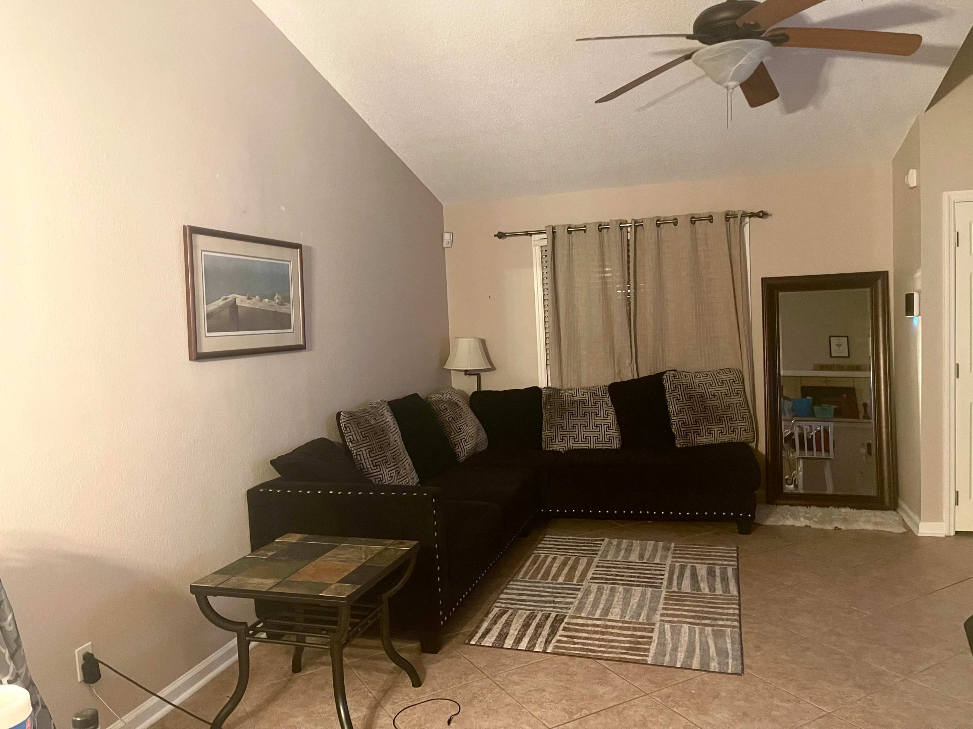 A living room with a dark sectional sofa, a patterned area rug, a small table, and a ceiling fan.