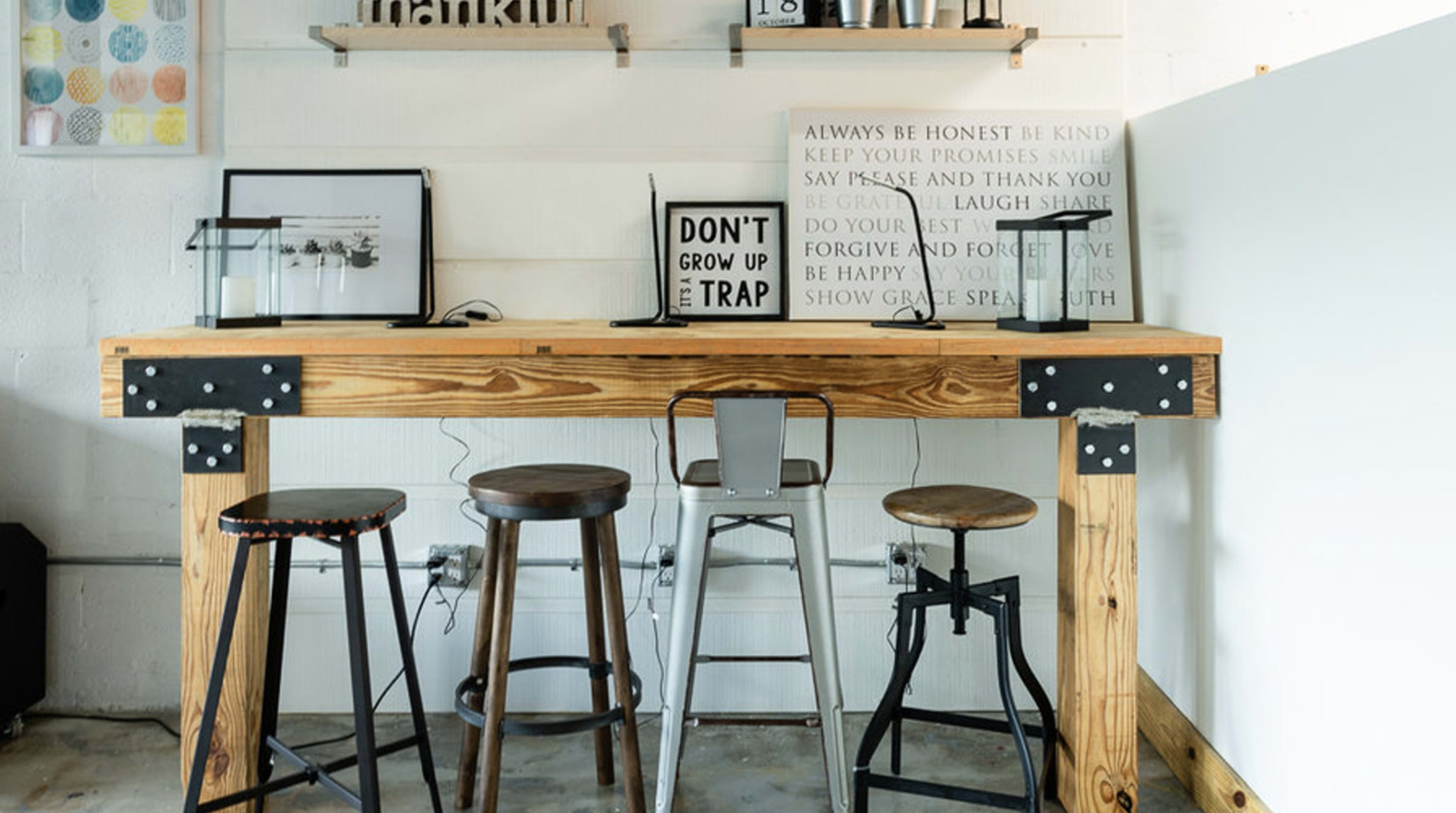 A wooden bar table with a metal frame and four stools arranged around it, set against a wall with various decorative items and framed signs.