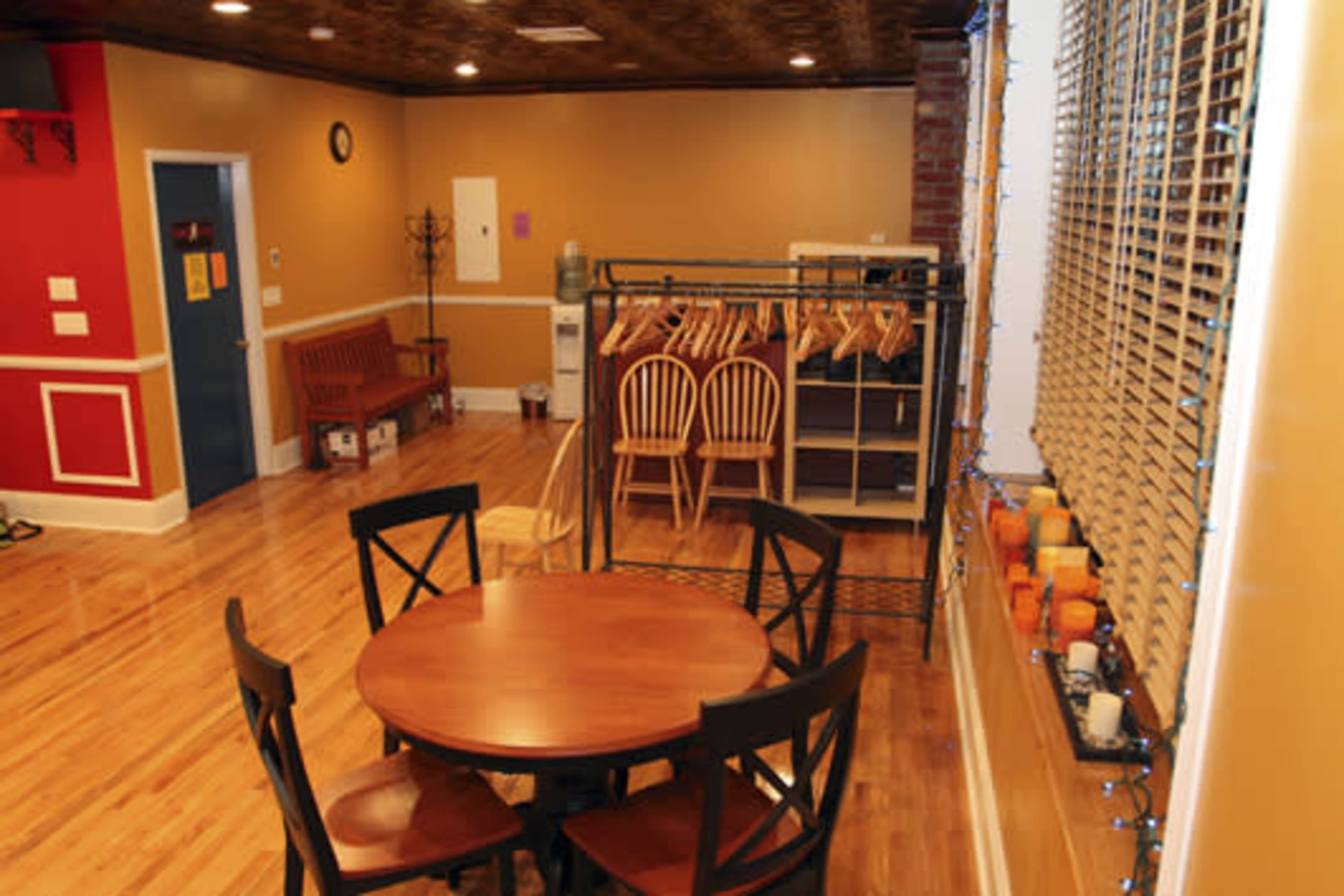 The image shows a brightly lit room with wooden flooring, featuring a round dining table with four chairs, a coat rack, and a section with hangers and various decorative items on a windowsill.