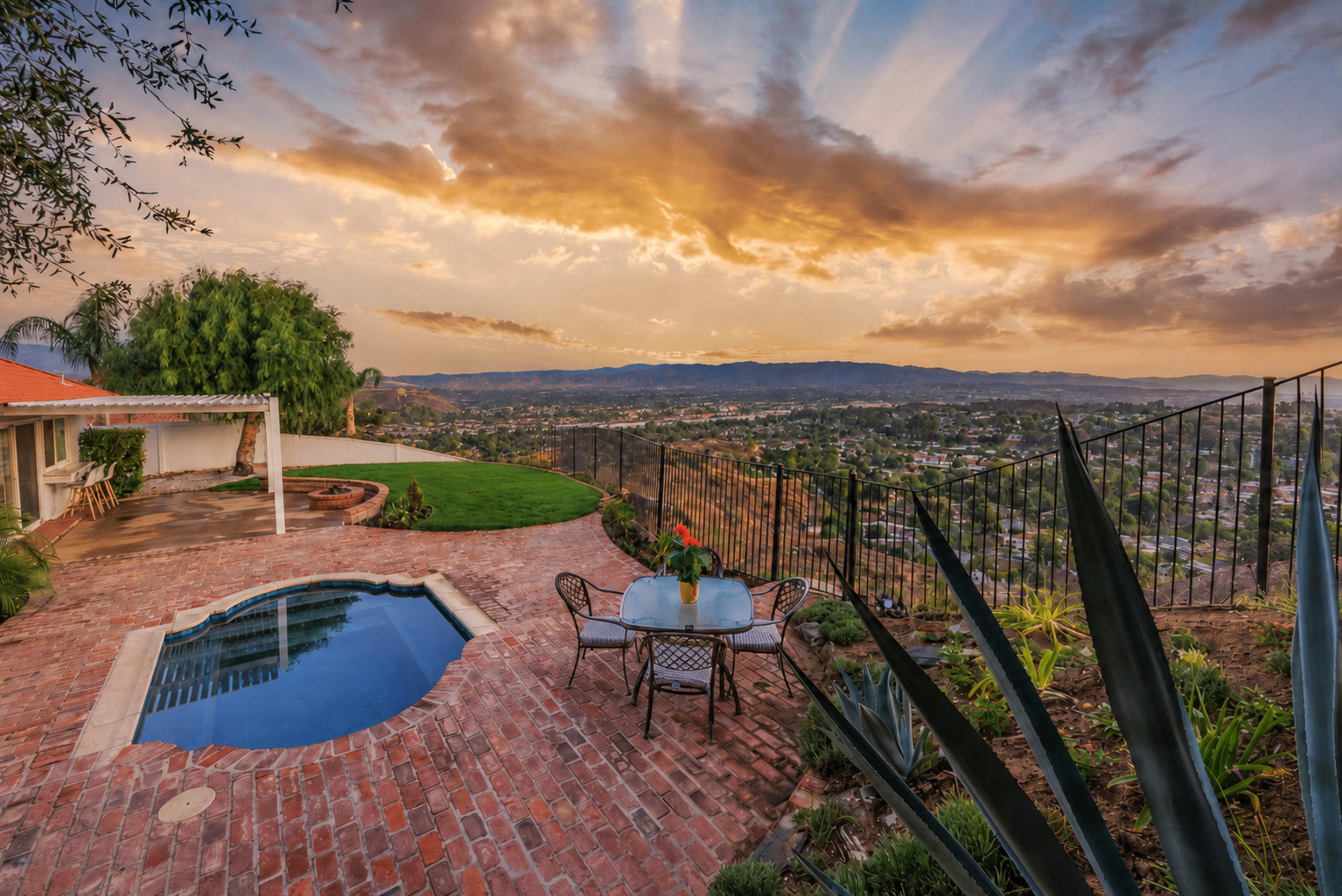 A small patio with a pool overlooks a valley at sunset, featuring a table set for two with a pitcher and glass.