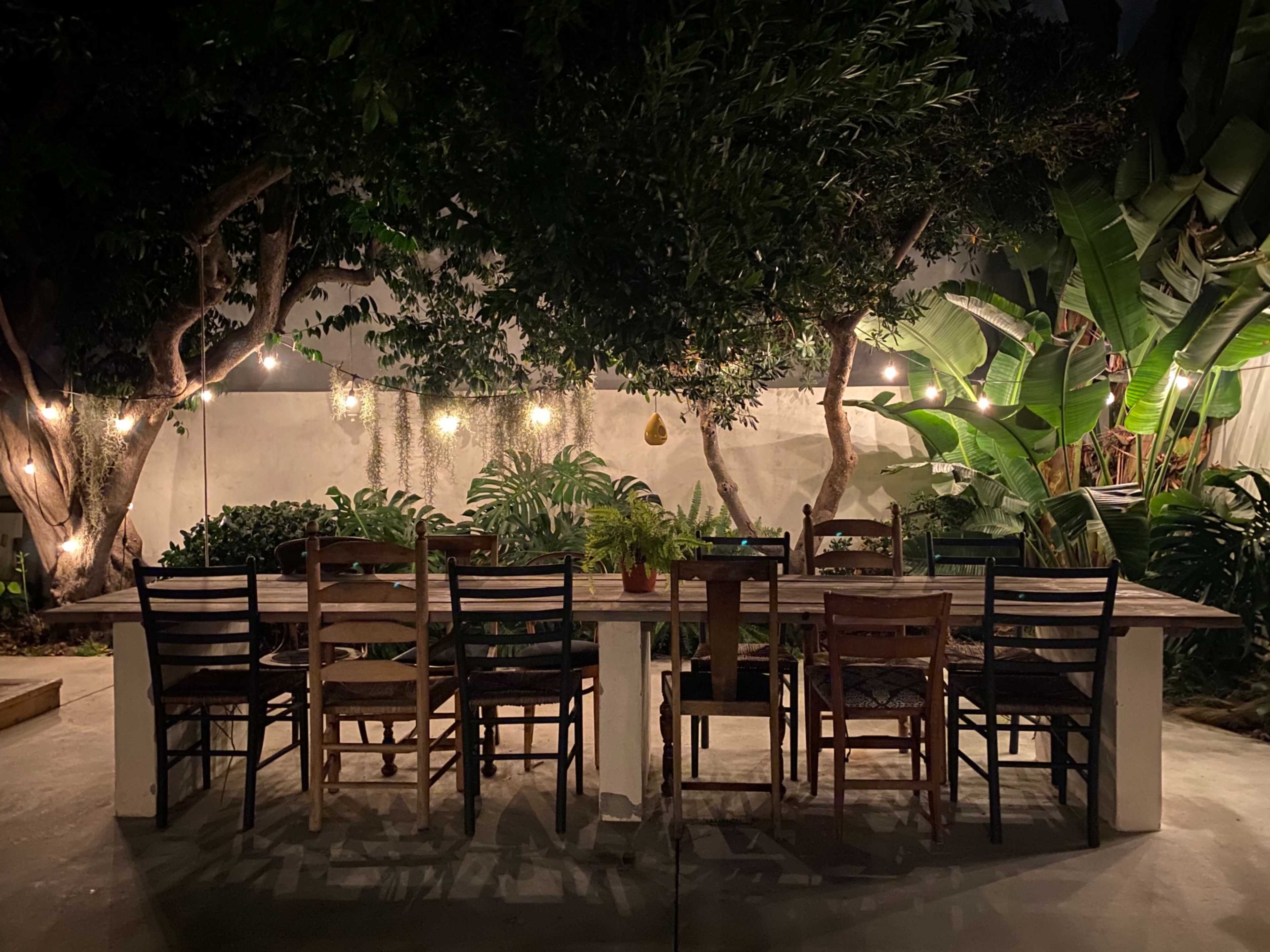 A long dining table with multiple chairs is set under string lights and surrounded by lush greenery.