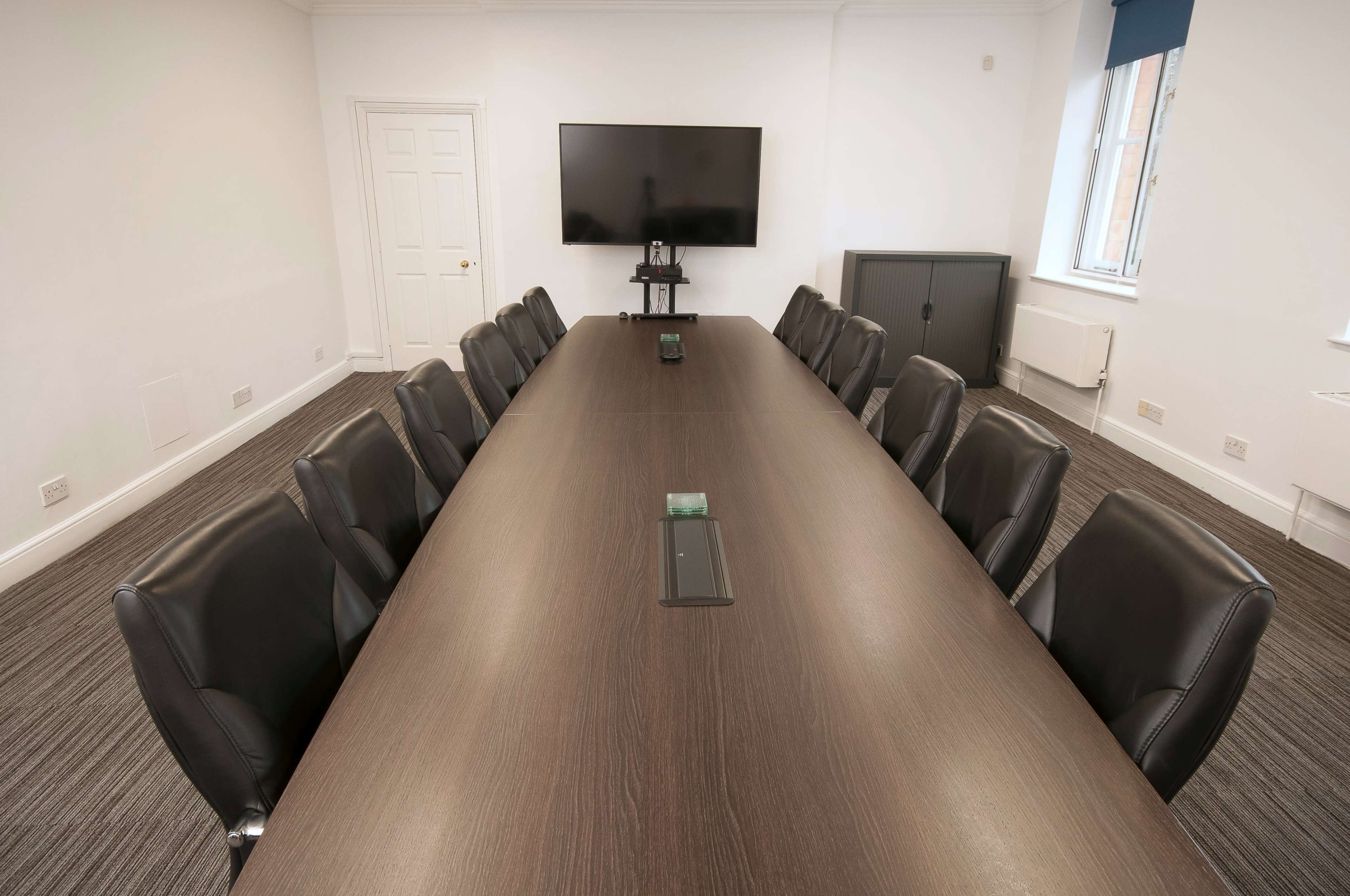 A large conference room features a long wooden table surrounded by black leather chairs, with a television mounted on the wall.