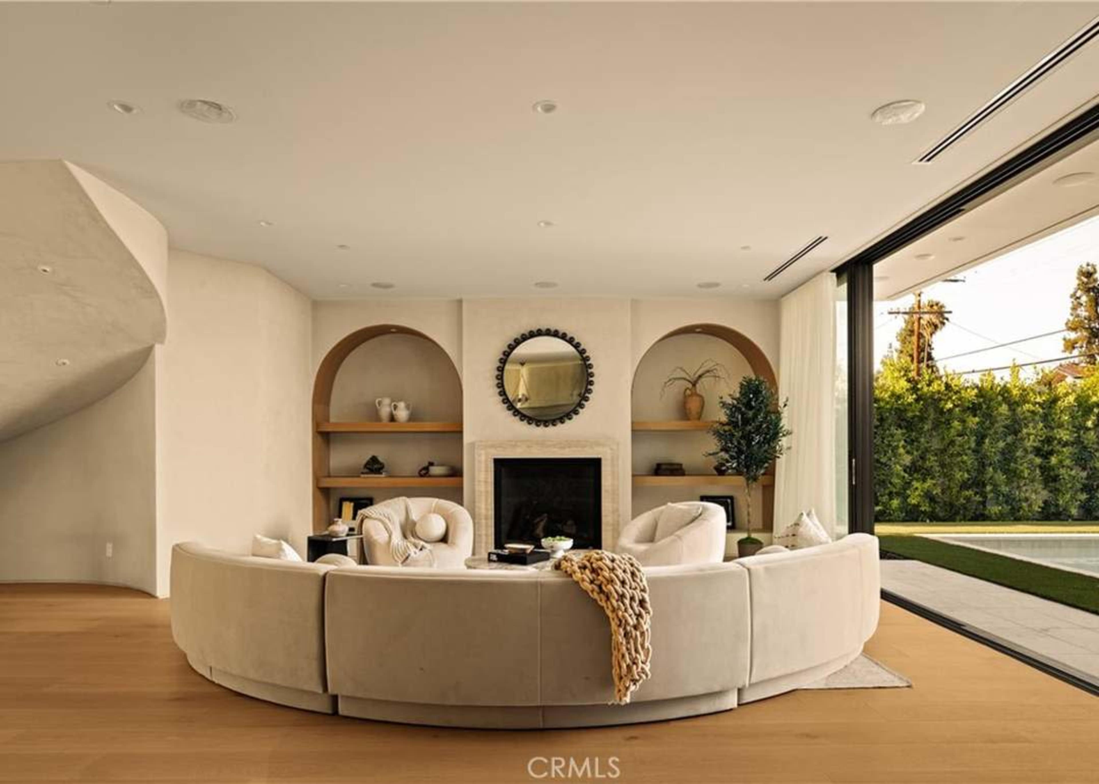 A circular white sofa is arranged in a modern living room with a fireplace, built-in shelves, and large sliding glass doors leading to an outdoor area.
